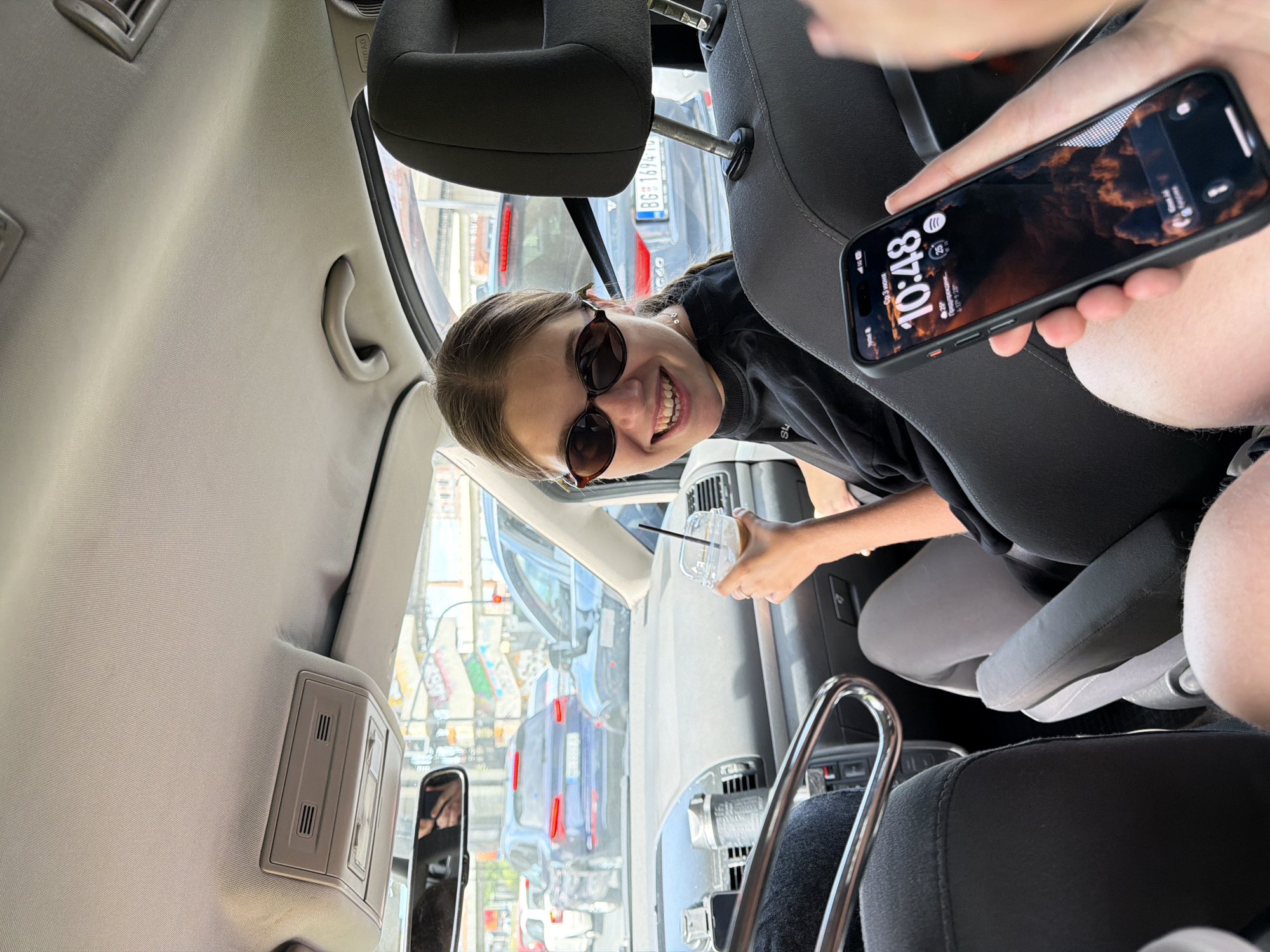 An interior shot of a car shows a young woman in the front passenger seat smiling towards the camera, while another person in the foreground holds a smartphone. The image is taken from a low angle, likely from a back seat, looking towards the front. The young woman is wearing sunglasses and a dark t-shirt, holding a clear plastic cup with a straw.
Through the car's windows, other vehicles are visible, suggesting the car is on a road or in traffic. A Serbian license plate "BG 1694-TY" is clearly visible on a black car in the background, confirming the location as Belgrade, Serbia. The car interior features light-colored headliner fabric, a grab handle, air vents, and part of the rearview mirror on the left. Text "AIRBAG" is visible on the upper right pillar of the car.
The person in the foreground holds a black smartphone displaying the time "10:48". Other text on the phone screen, in Serbian, indicates "Понедељак" (Monday), "3. септембар" (September 3rd), and weather information: "25°" (25 degrees Celsius) and "Облачно" (Cloudy). The young woman appears cheerful, and the overall impression is of a casual moment captured while traveling in a car.