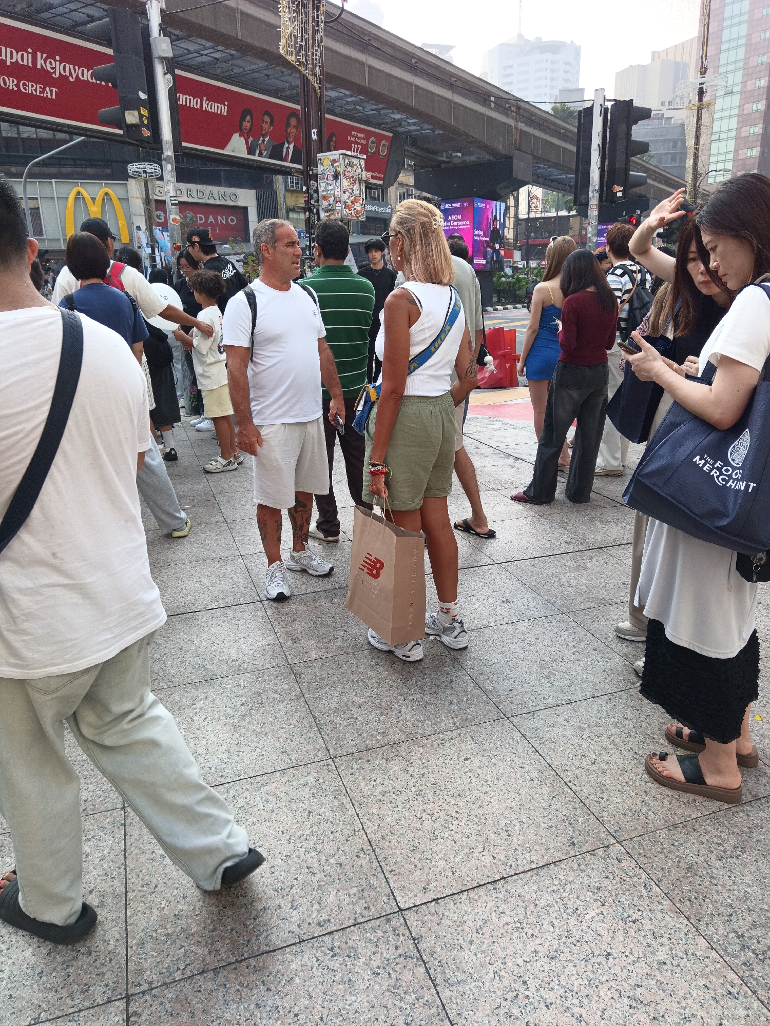 The image captures a street scene in Kuala Lumpur, Malaysia, likely near a commercial area given the presence of prominent signage such as "Apa Kejayaa..." and "ama kami" on a large red billboard overhead, along with a McDonald's logo. The ground is paved with textured tiles. A group of people are standing and walking on the pedestrian walkway.
In the foreground, a man with tattoos on his legs, wearing a white t-shirt and light-colored shorts, stands facing away from the camera. To his left, a person in a white t-shirt with a black strap across their chest is mostly visible from the back. To the right of the man with tattoos, a woman with blonde hair, wearing a white tank top and olive green shorts, holds a brown paper shopping bag from New Balance. Another woman in a blue dress is visible behind her.
Further in the background, other individuals are present, including a man in a green and white striped shirt, and others dressed in casual attire. Some people appear to be looking at their phones or interacting with each other. The environment suggests a busy urban setting, possibly a shopping district, with a skybridge or elevated train track visible in the upper part of the image, common in Kuala Lumpur's urban landscape. The overall atmosphere is that of a public space with moderate pedestrian traffic.