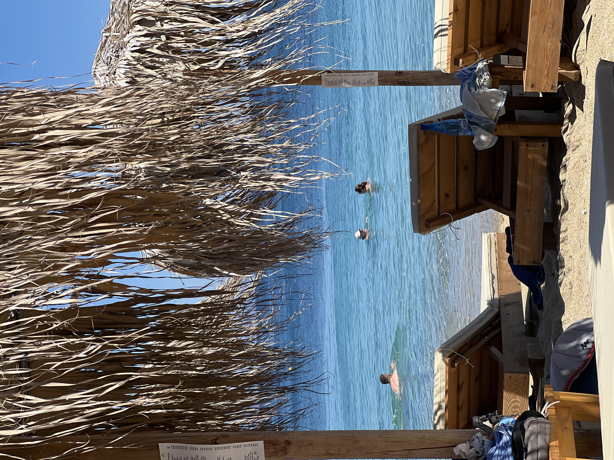 This image depicts a beach scene in Achladias, Greece. In the foreground, two wooden lounge chairs are partially visible on the sand, covered by a thatched umbrella. Beyond the chairs, the clear blue water of the sea stretches to the horizon. Three individuals are in the water: one man is swimming further out, while two people, one wearing a hat, are closer to the shore. The sun is shining brightly, casting shadows on the sand and illuminating the water. A menu is attached to a wooden post supporting the umbrella on the left side of the image.