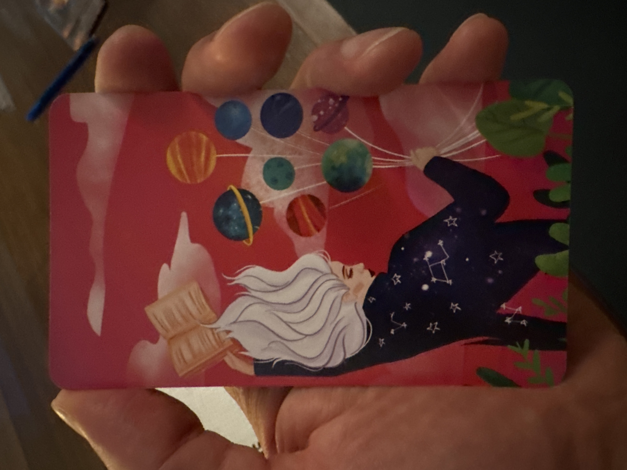 A playing card with an illustration is held in a human hand. The illustration features a woman with white hair lying on her side, wearing a dark blue shirt or jacket decorated with stars, resembling a constellation pattern. The background of the card is predominantly pink with white cloud-like shapes. The woman is depicted holding strings attached to several planets, which appear as colorful balloons. An open book rests near the woman's head. Green foliage is visible on the right side of the card. There is no indication this is located in Moscow, Russia.