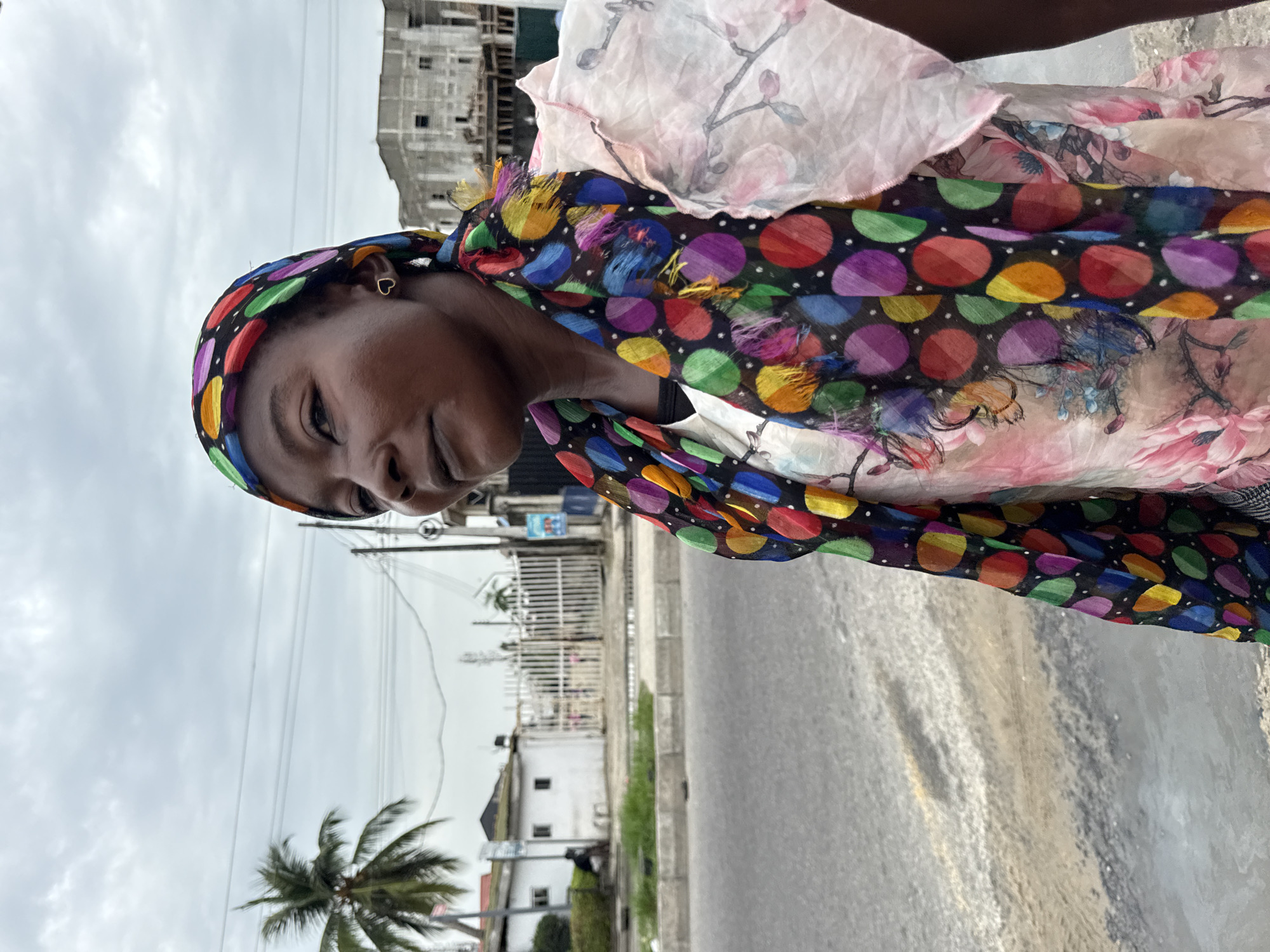 A young Black woman is outdoors, facing the camera and slightly left. She is wearing a headscarf with a black background and colorful polka dots. A floral print scarf is layered on top. She has heart-shaped earrings. The background shows a street scene with buildings, a palm tree, utility poles and wires, and a cloudy sky. The location is urban, possibly Ado, Nigeria. The scene appears to be a street portrait with the buildings and palm trees indicating the urban environment and location.