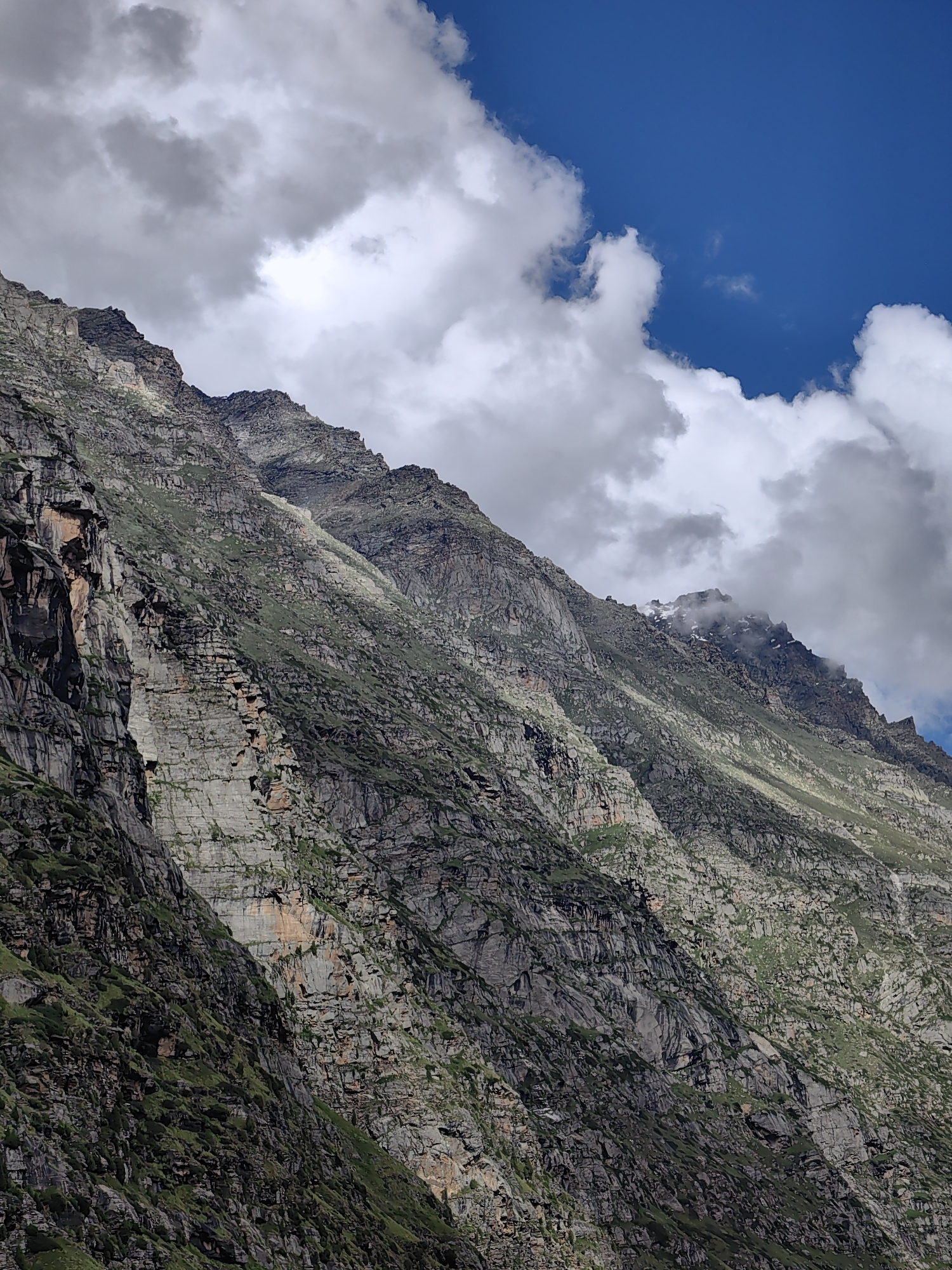 The image shows a steep, rocky mountain range under a blue sky with scattered clouds. The mountains are covered in patches of green vegetation and show signs of erosion and rockfall. The texture of the rock is rough and uneven, with visible strata and fissures. The lighting suggests it is daytime, with some areas of the mountain in shadow and others illuminated by sunlight. The overall impression is one of a rugged and natural landscape. The location is identified as Unknown, India.
