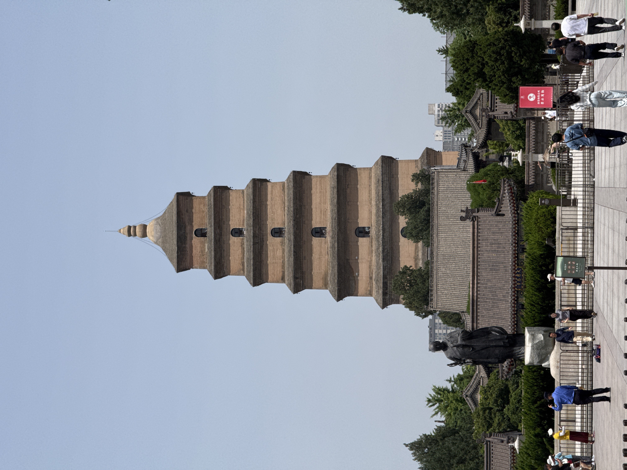 The image shows the Giant Wild Goose Pagoda, a large Buddhist pagoda located in southern Xi'an, Yanta District, China. It stands tall against a clear, pale blue sky. The pagoda is made of brick and has a distinctive tiered design, with seven layers that taper as the structure ascends. Small, dark rectangular windows are visible on each tier.
The lower part of the image shows a section of the surrounding grounds. A statue of a figure, likely a historical or religious figure, stands on a platform with people walking nearby. Some are facing the camera and others are turning away. The grounds include pathways, fencing, landscaping with trees, and traditional Chinese-style buildings with tiled roofs. A red sign with text is seen in the background.