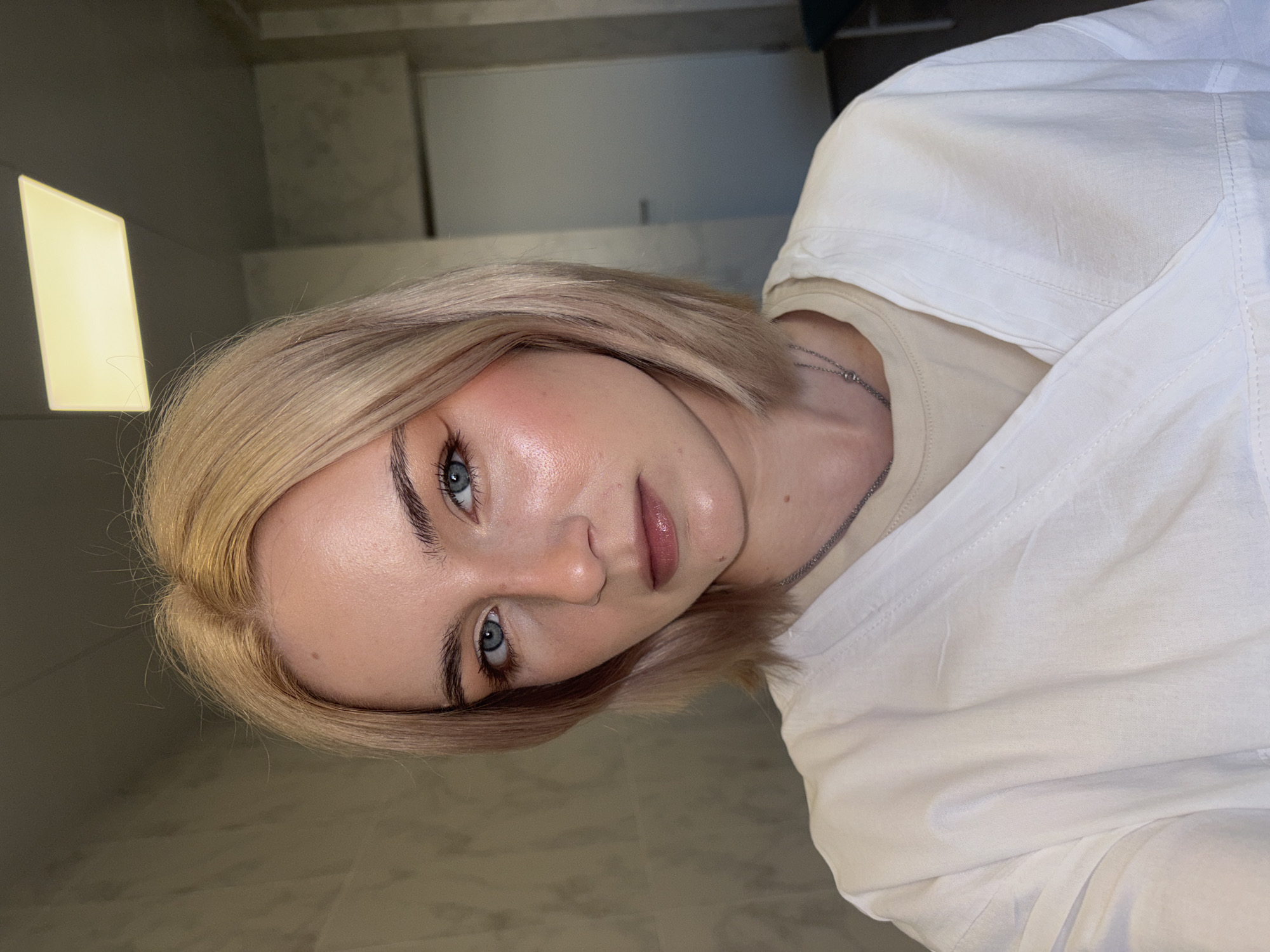 A selfie captures a young woman indoors, looking directly at the camera with a neutral expression. She has short, straight blonde hair and striking blue eyes, complemented by well-defined dark eyebrows. Her fair skin shows a subtle blush or highlight on her cheekbones, and her lips are painted with a soft pinkish-brown lipstick. She is wearing a light-colored, possibly white, collared shirt or jacket over a beige or tan round-neck top. A thin silver chain necklace is visible around her neck.
The background is indistinct, appearing to be an indoor setting with plain walls and some architectural lines or shadows. To the left, a rectangular light fixture emits a warm glow. The image is rotated 90 degrees clockwise, placing the woman's head tilted to the left. The lighting is soft but bright, highlighting her facial features. No text is visible in the image. The location context is unknown, Russia.