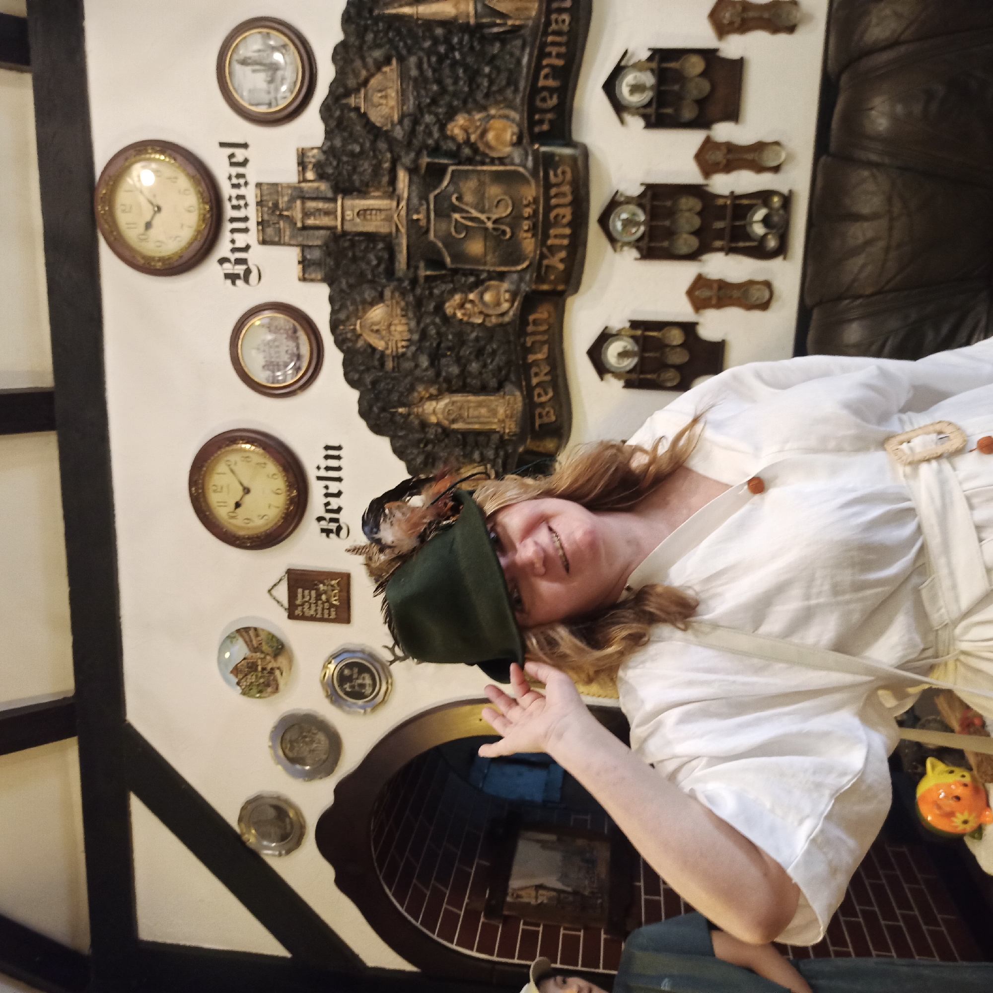 A medium shot captures a woman wearing a white dress and a dark green hat adorned with feathers. She stands indoors, with a wall decorated with various items behind her. These include several round clocks, small decorative plates, and a large, dark relief sculpture depicting buildings, possibly landmarks, with the words "Berlin" and "Brussel" visible. The sculpture also has text that reads "1995 Knaus" and what appears to be Cyrillic lettering "ЧЕРНІВЦІ", which transliterates to "Chernivtsi". Below the large sculpture, several small decorative clocks are mounted on the wall. To the left of the woman, a part of another person wearing a green garment is visible. The environment appears to be a themed interior, possibly a restaurant or museum, given the collection of clocks and decorative items. The overall lighting suggests an indoor setting.
