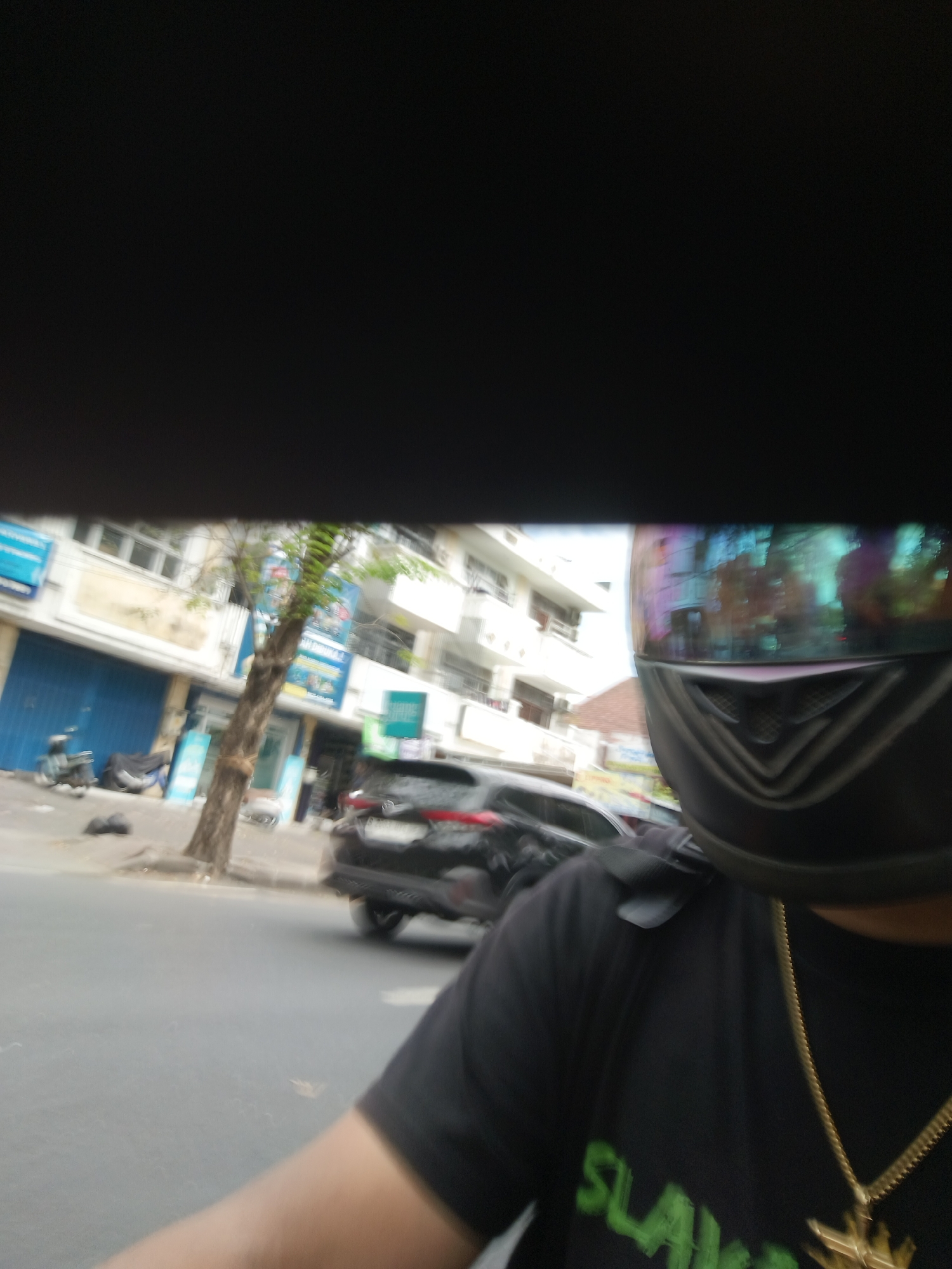 The image is a blurry, first-person perspective of someone riding a motorcycle in Denpasar, Indonesia. The rider is wearing a black t-shirt and a helmet with a colorful, reflective visor. In the background, there are buildings, a tree, and a black car. The street appears to be paved and there is some traffic. The overall impression is a candid snapshot of daily life in a busy urban environment.
