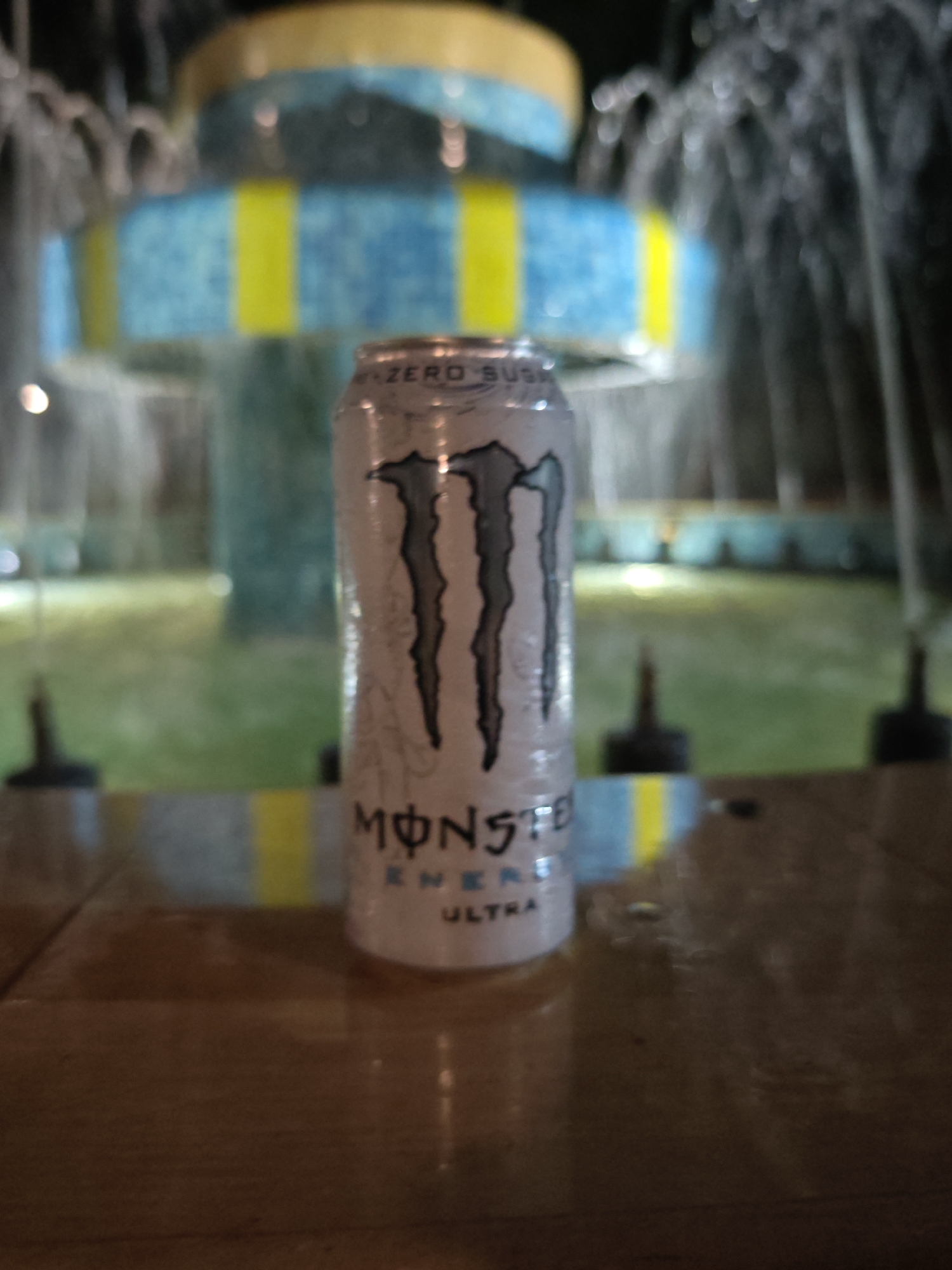 An upward-angled, eye-level photograph captures a single white Monster Energy Ultra can positioned centrally on a smooth, light brown surface, likely a table or ledge, in Veliki Preslav, Bulgaria. The can, exhibiting condensation, features the prominent black "M" claw logo and the visible text "MONSTER ENERGY ULTRA" and "ZERO SUGAR." The background is significantly out of focus, depicting a multi-tiered fountain illuminated by artificial light. The lower tier of the fountain is blue with distinct vertical yellow stripes, while an upper tier appears yellowish. Streams of water emanate from the fountain, creating a blurred, misty effect. Behind the fountain, indistinct green foliage or grass is visible, along with multiple small, dark, conical objects on the ground, possibly bollards or light fixtures. Blurred points of light, indicative of a low-light or nighttime environment, are scattered throughout the background. No individuals or discernible actions are present in the image.