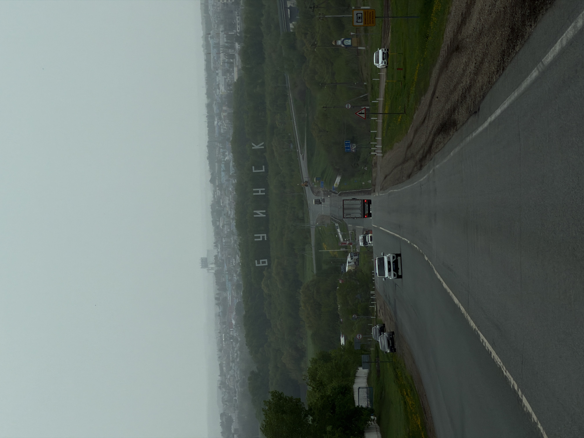 The image depicts an elevated view of a road leading towards the city of Minsk. The city's name, "МИНСК" ("MINSK" in Cyrillic), is spelled out in large letters along a wooded slope leading down towards the urban area. The foreground consists of the road, with several vehicles, including cars and a truck, visible. The weather appears overcast and hazy. The image suggests the view from a roadside overlook. The location is likely somewhere near Stepanovka, Russia.