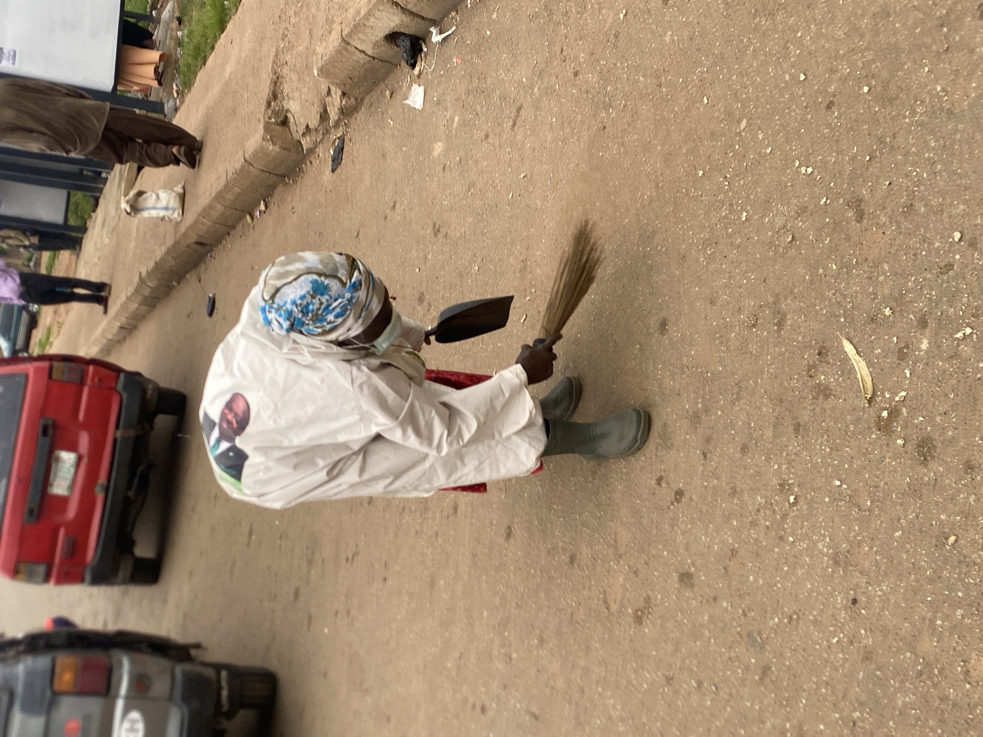 An individual, dressed in a white garment and a blue and white headscarf, is sweeping a dusty road in Bukuru, Nigeria. They are wearing green boots and a face mask. A dark dustpan is held in their right hand, while a broom is being used with their left hand. The person is bent over, actively engaged in sweeping. In the background, the rear of a red vehicle is visible. Several other individuals are standing on a sidewalk to the right, with a white bag placed on the curb. The environment appears to be an outdoor, urban setting with scattered debris on the road and sidewalk.