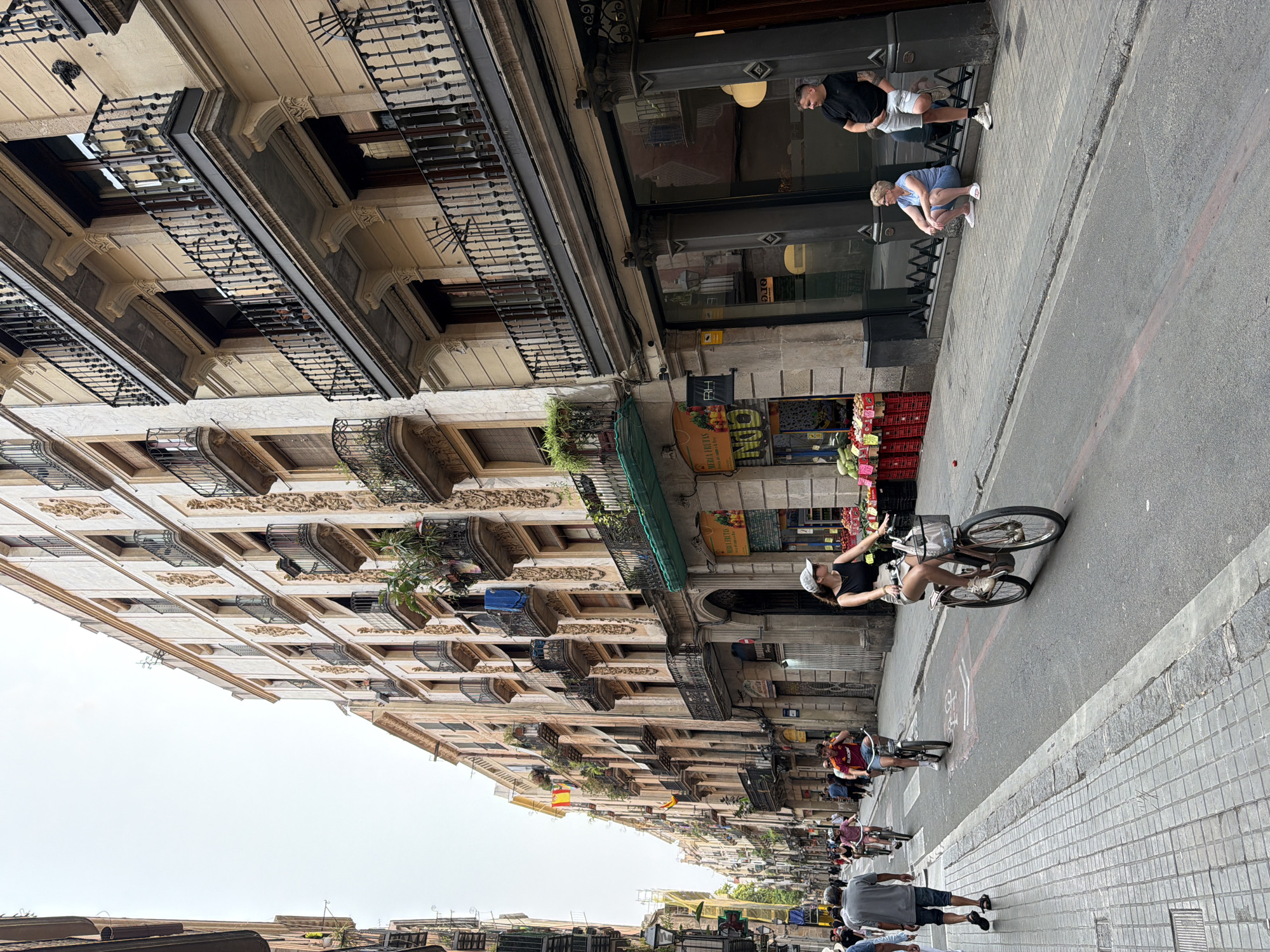 In Barcelona, Spain, a street scene unfolds with people navigating a narrow street lined with tall, ornate buildings. The architecture features multiple stories with balconies adorned with wrought-iron railings, and some balconies have plants. The street is paved with cobblestones on the sidewalks and asphalt on the road, which has painted lane markers and a designated bicycle lane.
Several individuals are on bicycles, including a woman in the foreground wearing a white hat and a dark top and shorts, riding a brown bicycle. Another person on a bicycle is further down the street. Pedestrians are also present, with one man in a grey shirt and shorts walking on the sidewalk.
To the right, a storefront displays fruits and vegetables. Two people are near this storefront: a woman is sitting on the curb wearing a blue top and shorts, and a man in a black t-shirt and light shorts is standing with his arm resting on a pillar.
The sky is overcast, suggesting a cloudy day. The overall impression is of a lively urban environment in Barcelona with a mix of residential and commercial activity.