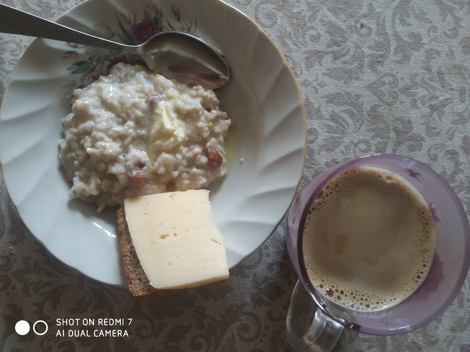 A top-down medium shot depicts a meal with a beverage, likely photographed in the city of Chaikovsky, Russia. The meal is placed on a table covered with a patterned tablecloth. A white bowl with a floral design holds cooked oatmeal with pieces of butter melted into it. A piece of cheese rests on a small slice of dark bread next to the oatmeal. A metal spoon lies within the bowl. To the right of the bowl, there's a glass mug filled with a tan-colored beverage, potentially coffee with milk or cappuccino, which has visible bubbles on the surface. A "SHOT ON REDMI 7 AI DUAL CAMERA" watermark is at the bottom left of the image. The environment is simple, focusing primarily on the breakfast set-up.