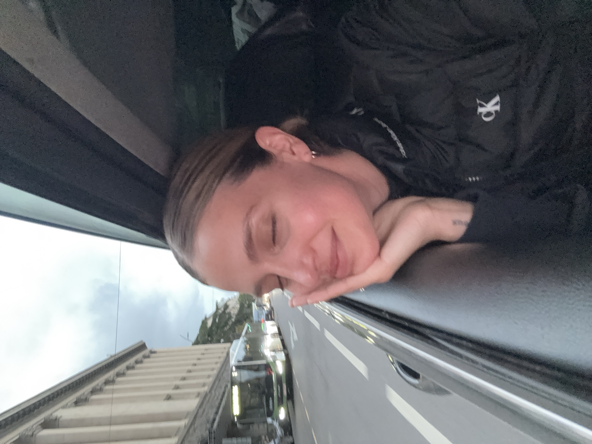 The image captures a serene moment of a young woman resting inside a vehicle, likely a bus or car, with her chin propped on her right hand and her eyes gently closed. Her hair is pulled back, revealing a small earring. She is wearing a black puffer jacket, prominently featuring a white "CK" logo, indicating Calvin Klein. A faint, unreadable tattoo or text is visible on her right hand or wrist.
Outside the window, an urban street scene unfolds under an overcast or cloudy sky, suggesting daytime without direct sunlight. To the left, a distinctive building or structure with a ribbed or stepped facade is visible. A road with white lane markings stretches ahead, where a dark-colored bus with its lights illuminated, along with other vehicles, can be seen. The overall atmosphere conveys a peaceful moment of travel or contemplation. The scene is set in Tbilisi, Georgia.