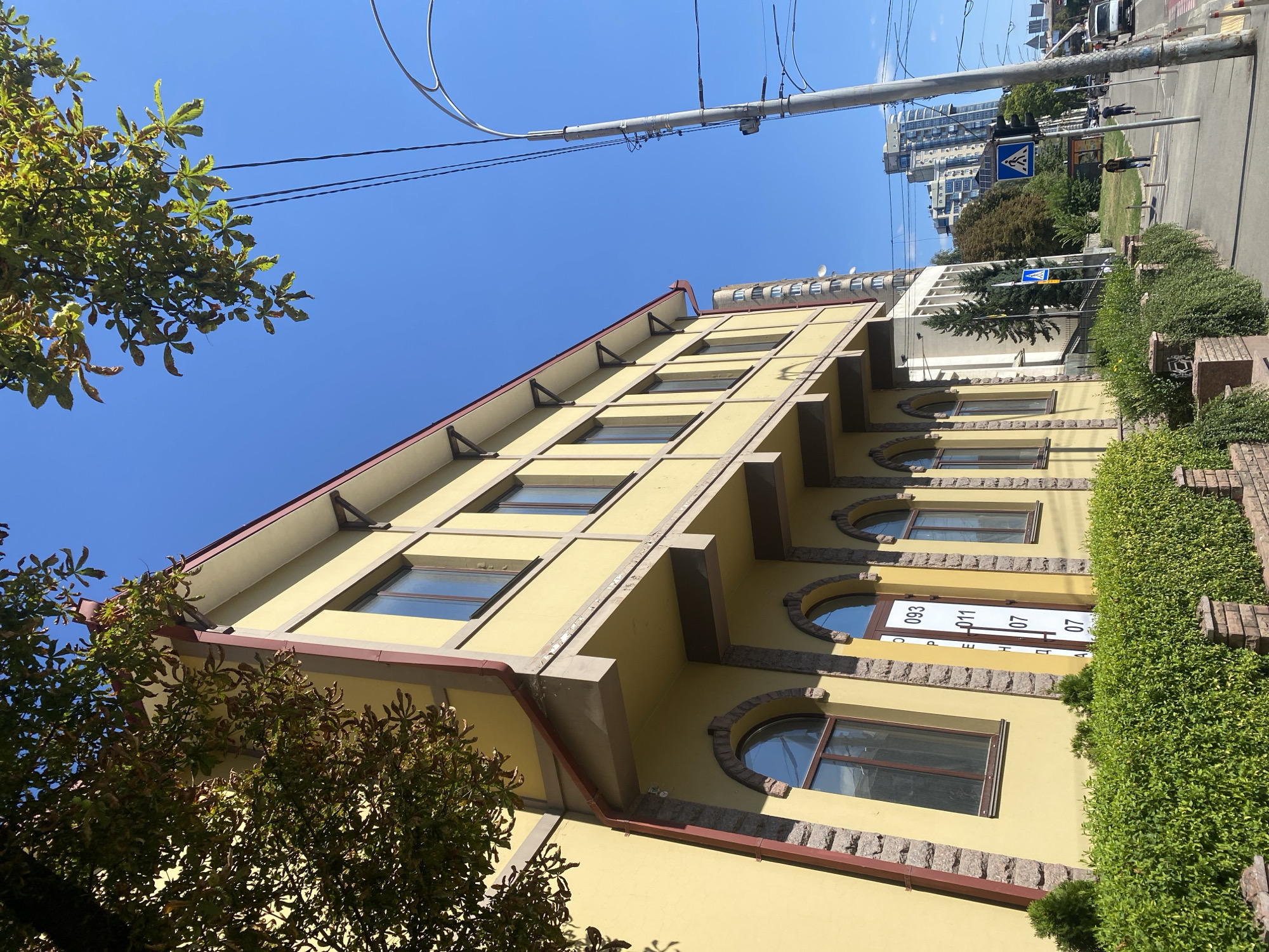 The image depicts a yellow, multi-story building with a burgundy trim, viewed from a low angle. The building features arched windows on the lower level, framed by stone accents, and rectangular windows on the upper level. A dense green hedge runs along the front of the building, with brick pillars interspersed. The sky is clear and blue. To the right of the building, a sidewalk runs alongside a street. A lamppost with electrical wires is prominent in the foreground. Further down the street, there are several pedestrian crossings, traffic lights, and other vehicles. In the background, tall, modern buildings and trees are visible, suggesting an urban environment.