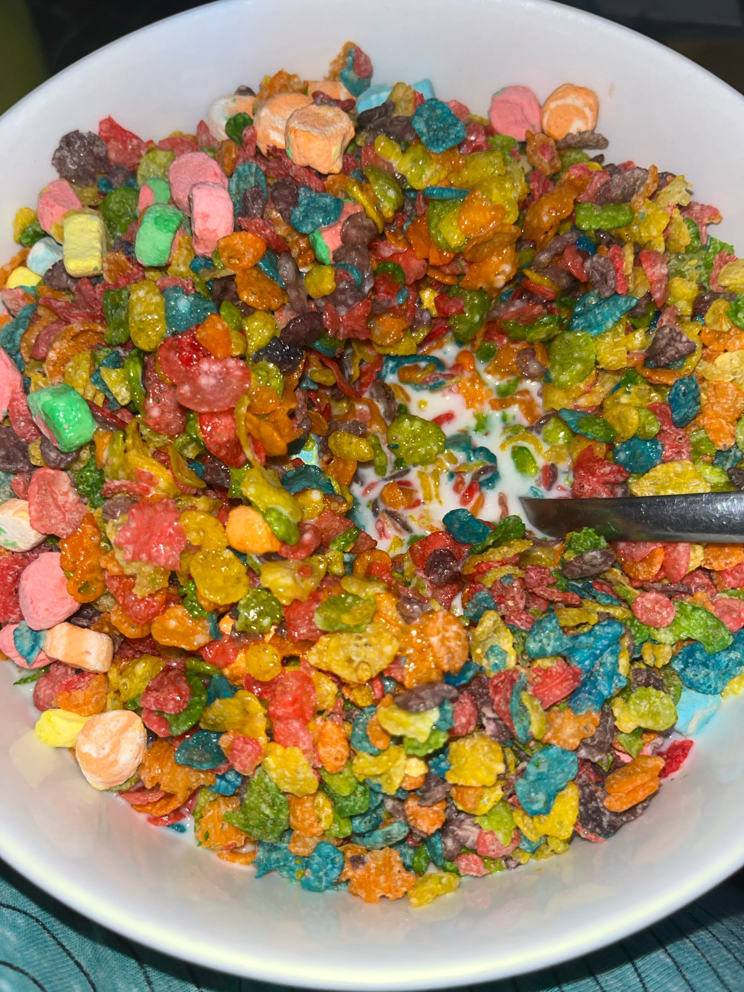 A close-up, overhead shot captures a white bowl filled with a colorful assortment of fruity, marshmallow-laden cereal and milk. A silver spoon rests partially submerged in the milk, angled towards the right side of the frame. The cereal exhibits a vibrant mix of red, orange, yellow, green, blue, and purple hues. The surrounding environment is not visible, and the image does not provide any information about a specific location in San Antonio, United States of America.