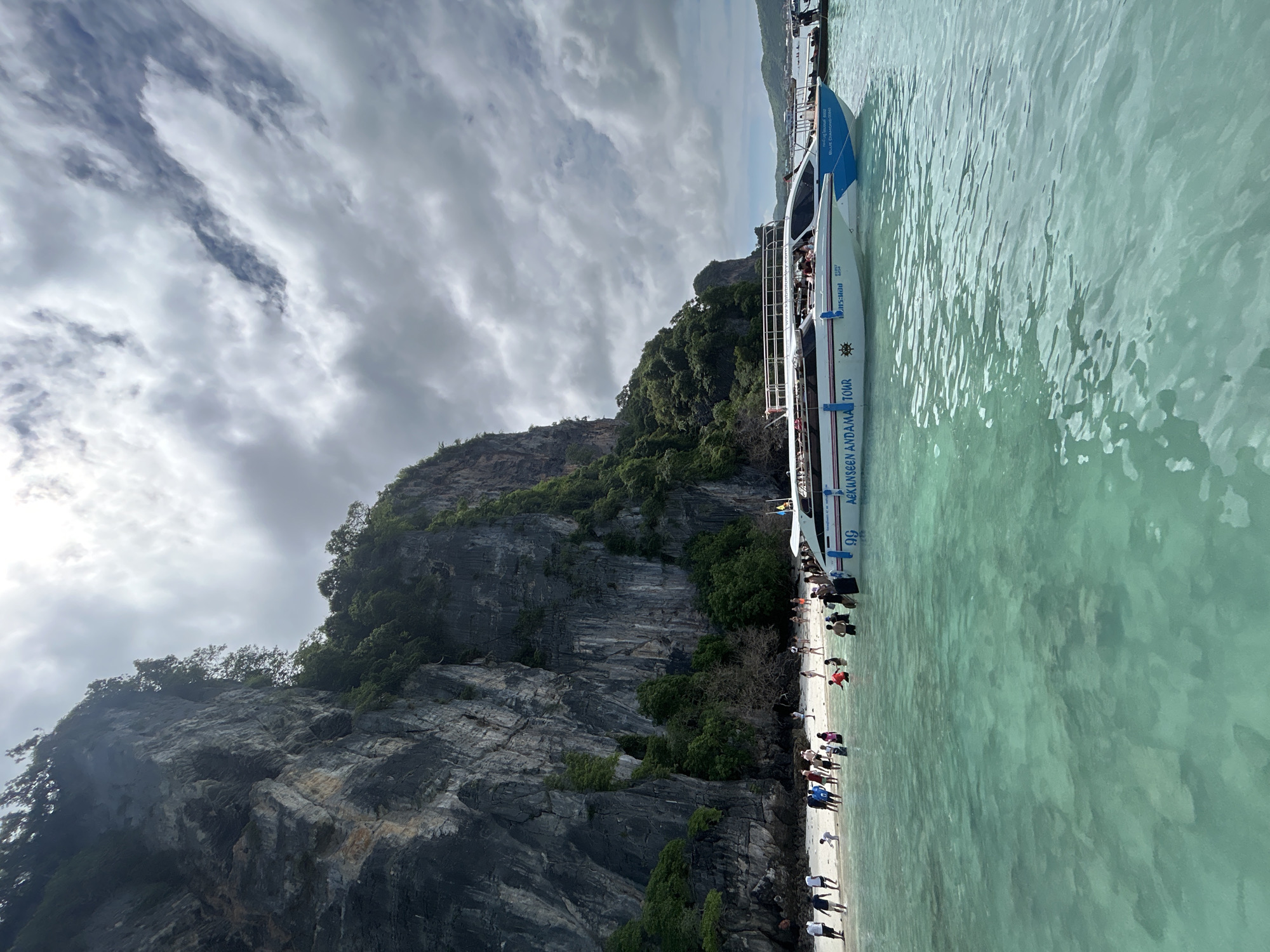 This image depicts a scenic view of a tropical beach with a steep, vegetated cliff face rising behind it. The water in the foreground is a clear turquoise, showing ripples on its surface. A white and blue speed boat, labeled "UNSEEN ANDAMAN TOUR," is anchored close to the shore. Numerous people are visible on the sandy beach and wading in the shallow water. The sky is overcast with dramatic clouds. The location is identified as Ko Phi Phi, Krabi Province, Thailand, known for its limestone karst formations and beautiful beaches.