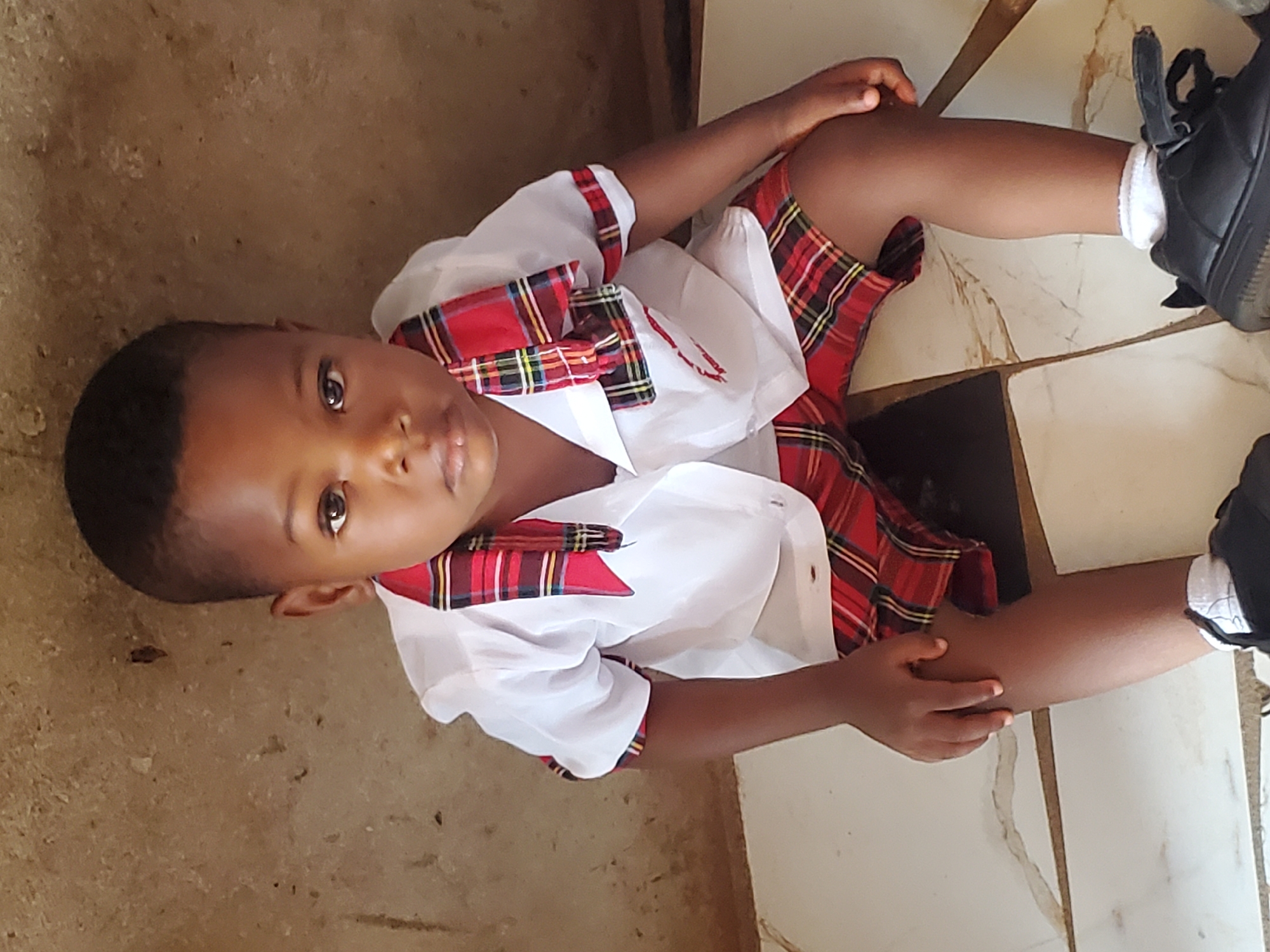 A young child is sitting on a tiled floor, possibly indoors. The child is wearing a white shirt with a red and black plaid trim, and matching plaid shorts. The child is wearing black shoes with white socks. The background consists of the tiled floor and a section of a wall. The location is inferred to be in Ukwu-Abwa, Nigeria.
