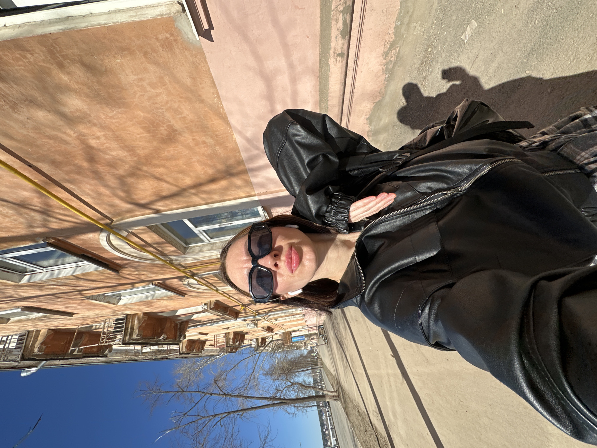 A woman stands on a concrete sidewalk in Votkinsk, Russia, on a sunny day. She wears a black leather jacket, dark sunglasses, and white earbuds. Her right arm is crossed over her chest. Behind her, a multi-story apartment building with visible window frames and utility pipes lines the street. Leafless trees border the sidewalk along with some bare buildings along a river. The sky is clear and blue. The time of year is probably either Spring or Fall.