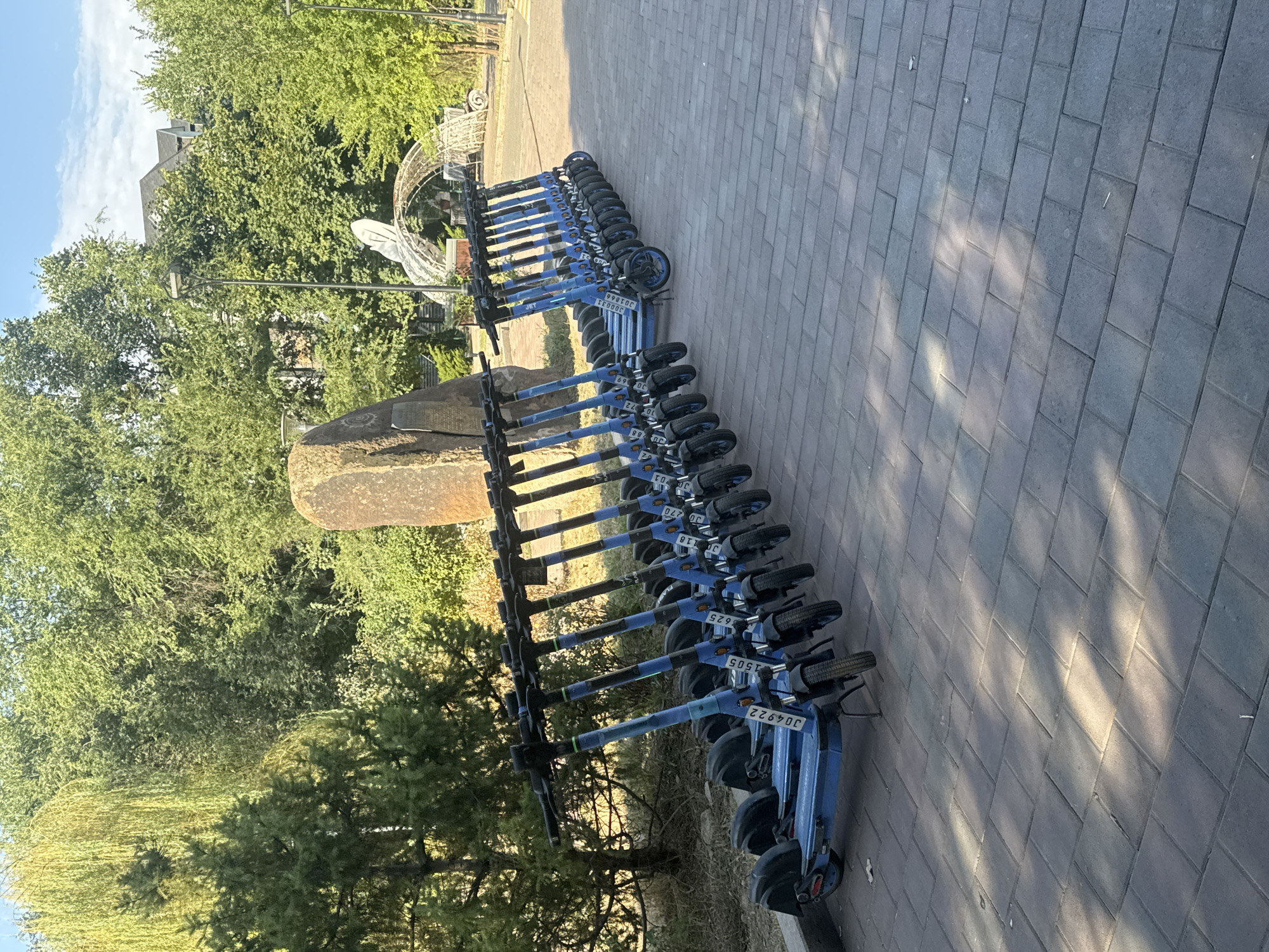 A row of blue electric scooters is parked on a paved walkway outdoors, with trees and a building in the background. The scooters are lined up neatly, with their handlebars facing forward. Some of the scooters have visible identification numbers. The walkway is made of rectangular paving stones. In the background, there are lush green trees, a large rock, and an architectural structure that appears to be a white archway. Sunlight casts shadows on the ground, indicating it is daytime. The scene suggests a public area or park. The image does not provide enough information to determine the specific location in Almaty, Kazakhstan.