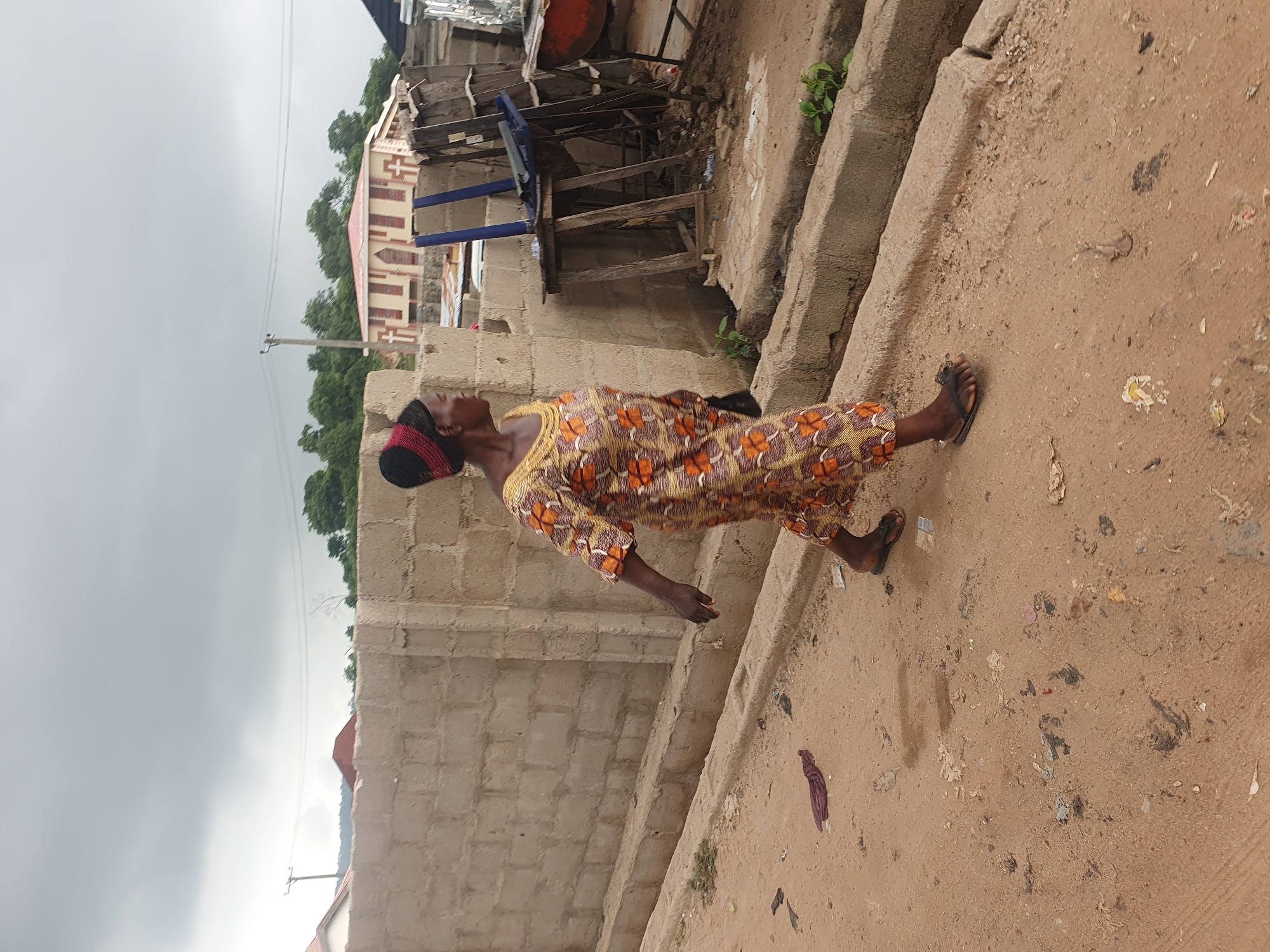 A woman with dark skin and dark hair, wearing a colorful African print dress and a red and black headwrap, walks on a dirt road. She is in profile, facing left. The dress has an orange, brown, and white geometric pattern. Her feet are bare except for dark flip-flops. She carries a dark bag in her left hand.
The environment appears to be an undeveloped area or outskirts of Lokoja, Nigeria. A rough, unpaved road is covered in sand and debris, including leaves, plastic, and other litter. A concrete drainage channel runs along the side of the road, with sparse green vegetation growing in it. Behind the woman, a wall constructed of concrete blocks stretches across the frame. In the background, there are more buildings with red roofs, some unfinished constructions, and utility poles with power lines. The sky is overcast and grey. The overall scene suggests a rural or semi-urban setting with some signs of construction and a lack of substantial infrastructure.