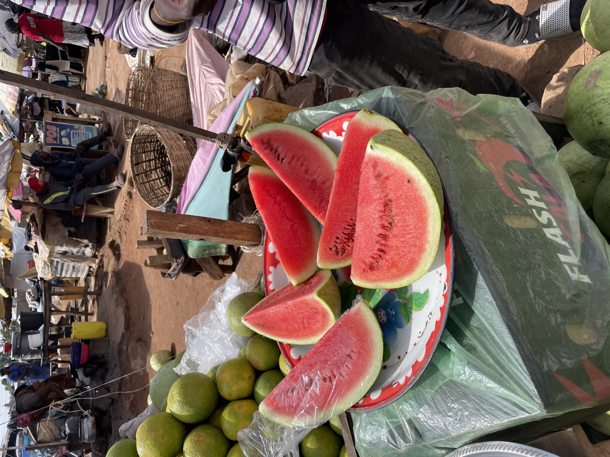 An outdoor market scene in Gura Top, Nigeria, displays various produce and bustling activity under bright daylight.
In the foreground, an assortment of fresh fruits is arranged for sale. Several vibrant red watermelon slices, some partially covered in clear plastic wrap, are neatly displayed on a decorative, circular metal tray with a red and white rim and blue floral patterns. Surrounding the tray are whole, light green watermelons. Adjacent to these, a large pile of round, uniformly green citrus fruits, possibly grapefruits or large oranges, rests on the ground. A large green plastic bag with the word "FLASH" prominently written in white capital letters, along with red graphic elements, is visible next to the watermelons.
Several people are present. A person wearing a distinctive purple and white striped shirt and dark trousers is positioned at the top of the frame, partially visible. In the background, two men are seated on a wooden structure, one wearing a red beanie and grey hoodie, the other in a dark hoodie. Other figures are less distinct in the busy background.
The market features rudimentary wooden stalls and structures, indicating an informal setting. Large woven baskets are visible, likely used for carrying or displaying goods. The ground is unpaved, consisting of reddish-brown earth. The bright sunlight casts distinct shadows, suggesting it is midday or early afternoon.
Visible text includes "POS" and "MTN" on signs in the background, and "VOICE" on a green container. The scene captures the essence of a lively local market.