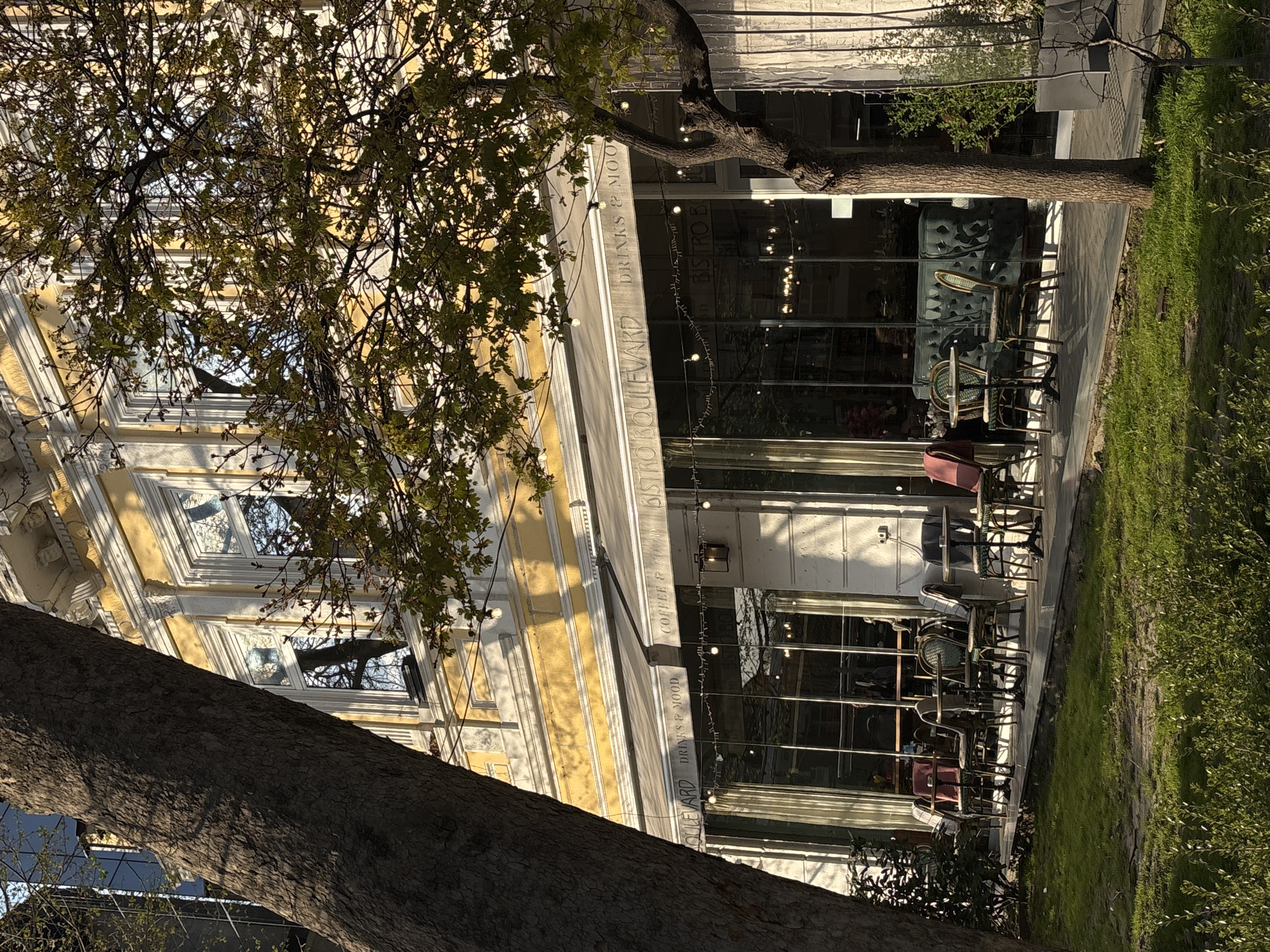 The image captures an outdoor scene featuring a cafe or bistro located in Varna, Bulgaria, seen from a slightly elevated perspective. The prominent building has an ornate, light yellow and white facade with classical architectural details and large, clear glass windows/doors. An awning extends over the entrance, and string lights are draped along the facade, suggesting a welcoming evening ambiance, though the current lighting indicates daytime.
On the building's facade and awning, text is visible: "BISTRO BOULEVARD", "DRINKS & MOOD", and "COFFEE & FOOD". Through the large glass windows, parts of the interior are visible, including a teal-colored sofa and internal lighting fixtures.
In front of the bistro, there is a paved outdoor seating area furnished with several tables and chairs, some of which appear stacked, possibly indicating that the establishment is not currently open or is preparing to open/close. To the right of the seating area, a patch of green grass and small bushes adds a natural element.
Two large trees with prominent trunks and branches, bearing light green leaves, partially frame the scene. One tree trunk diagonally crosses the lower left foreground, while another tree is closer to the building on the right side. The sunlight suggests either a bright morning or late afternoon, casting distinct shadows on the building and ground, contributing to the depth of the image. No people are visible, leaving the scene tranquil and focused on the architectural and natural elements.