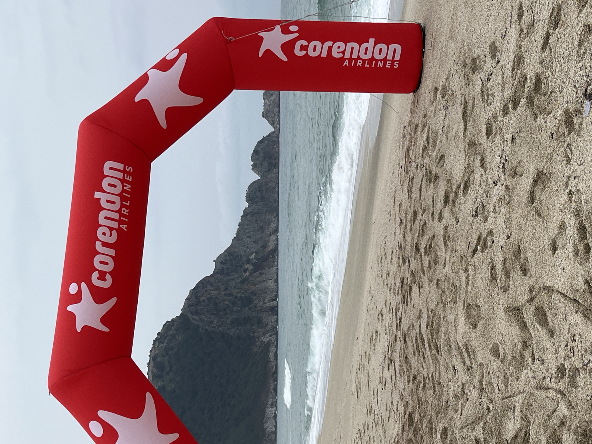 An outdoor scene captures a sandy beach in Alanya, Turkey. A prominent red inflatable arch, emblazoned with the white "Corendon Airlines" logo and a star-shaped emblem containing a human figure, stands on the beach. Anchoring ropes are visible securing the right support pillar of the arch. The foreground consists of light brown sand marked by numerous footprints. Beyond the sand, the Mediterranean Sea shows moderate waves breaking onto the shore. The background reveals a dark, rugged mountainous coastline with some green vegetation, extending into the sea. The sky is overcast. No individuals or active interactions are depicted.