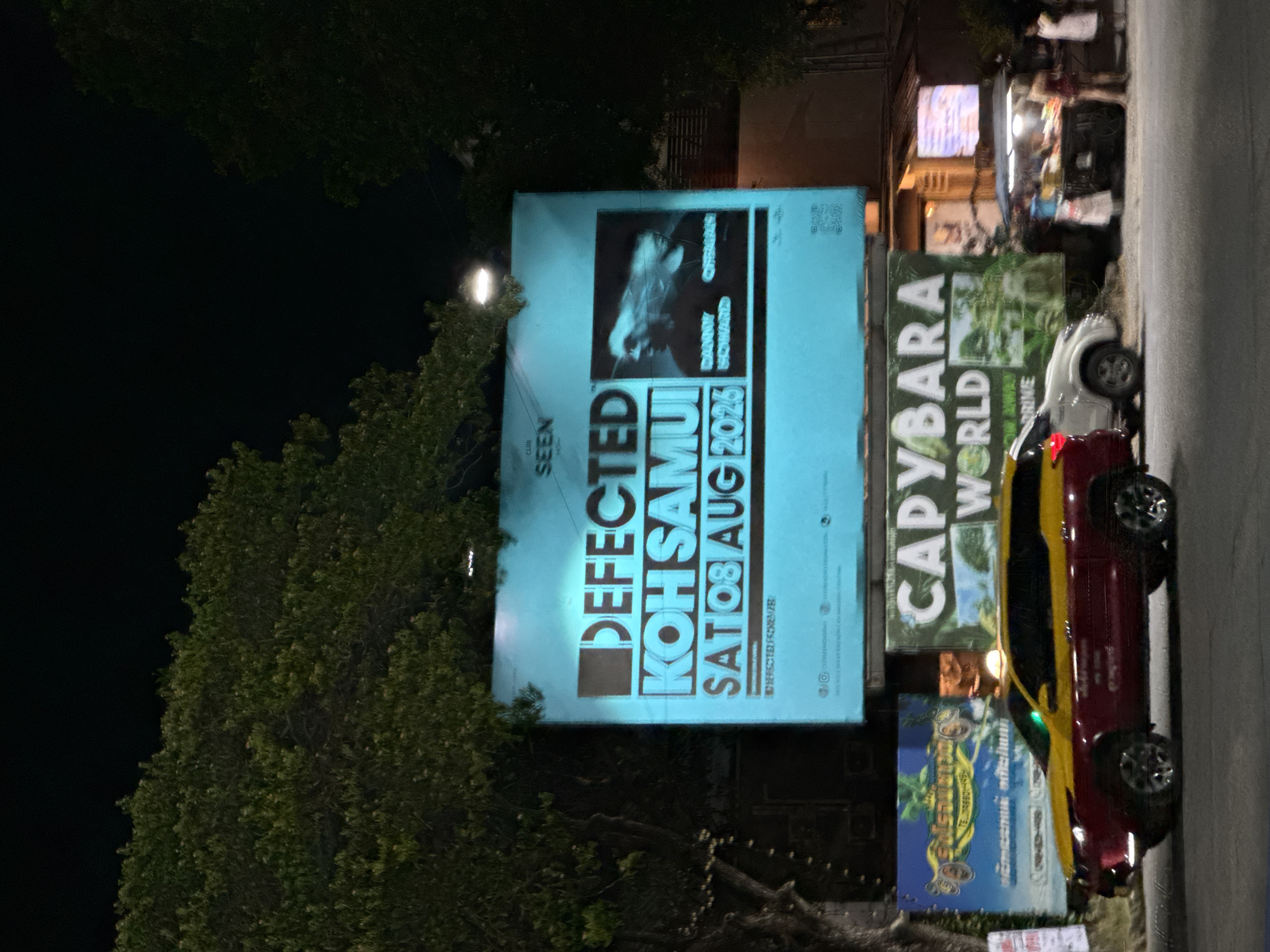 A nighttime, street-level, medium shot captures a vibrant scene in Koh Samui District, Thailand. Dominating the frame is a large, brightly lit turquoise billboard advertising "DEFECTED KOH SAMUI SAT 08 AUG 2025". The billboard features stylized text and a black-and-white image of two DJs, identified as "DANNY HOWARD" and "CHRISMAN".
Below the main billboard, a smaller, blue and yellow sign displays text in Thai script, partially obscured by a taxi. To the right of this, another billboard reads "CAPYBARA WORLD" with an image of lush greenery.
In the foreground, a distinctive two-toned taxi, red on the bottom and yellow on top, is parked on the street. To its right, a white SUV is visible. Further to the right, a street food stall is illuminated by a bright light, with several people gathered around it, appearing to be vendors and customers. The street is paved, and trees with dark foliage frame the top of the image, obscuring the night sky. The overall impression is that of a lively evening scene in a tropical urban environment.