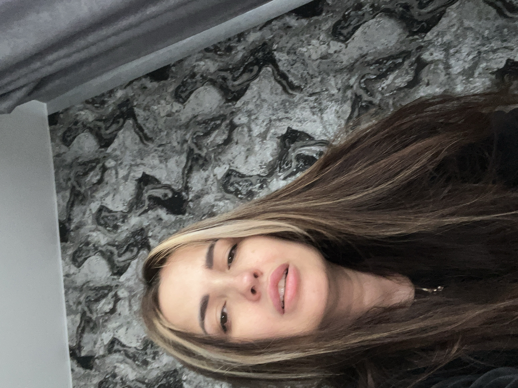 A head-and-shoulders portrait of a woman with long, brown and blonde highlighted hair is presented against a gray and black marble-patterned wall. A white wall or panel is visible on the left edge of the image. The woman's face is close to the camera, displaying her features, including her eyes, nose, and lips. The setting appears to be an interior space, with a gray curtain visible at the top of the image. The location is described as Unknown, Unknown.