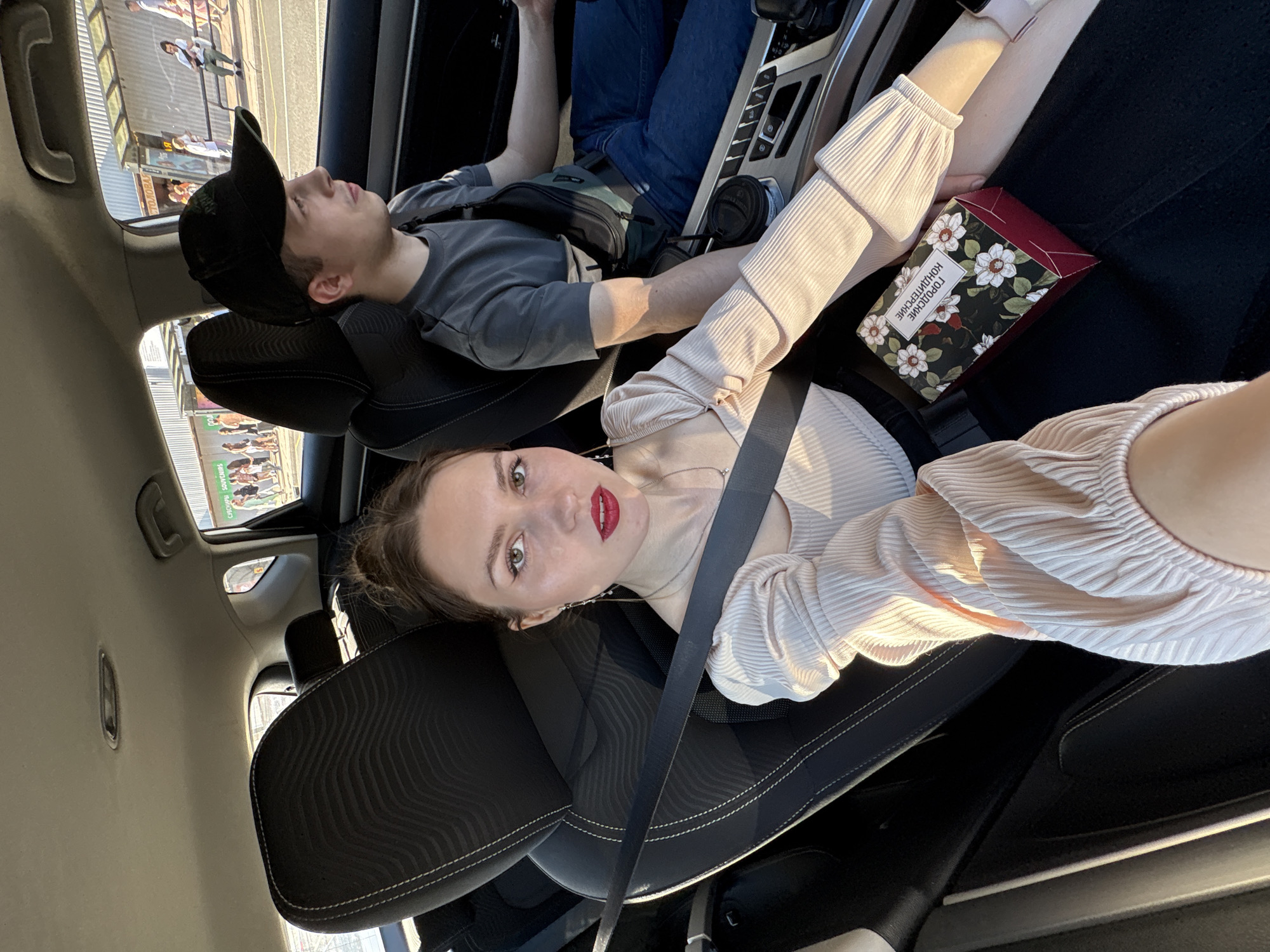 A selfie from the front passenger seat of a car captures a young adult female and a young adult male driving through an urban commercial street in Saint Petersburg, Russia, during daylight hours.
The female in the foreground, with pulled-back brown hair, red lipstick, and winged eyeliner, looks directly at the camera. She wears a light-colored, ribbed, long-sleeved top and a necklace with dangling earrings. A black seatbelt crosses her chest and lap. On her lap rests a dark-colored box labeled in Russian as "ГОРОДСКИЕ КОНДИТЕРСКИЕ" (City Confectioneries) with a white floral pattern.
To her right, the male driver is visible in profile, wearing a dark grey t-shirt, blue jeans, a black baseball cap, and a black cross-body bag. He looks forward, with a coffee cup in a holder within the center console beside him. He is also wearing a black seatbelt.
The car's interior features dark, textured seats with white stitching. Through the side windows, an active street scene is visible. Pedestrians walk on a sidewalk past low-rise commercial buildings. Signage includes "СУВЕНИРЫ SOUVENIRS" (Souvenirs) on a green banner and "СТОЛОВАЯ" (Canteen/Cafeteria) on another storefront. The architecture and street activity are consistent with a commercial district in Saint Petersburg.