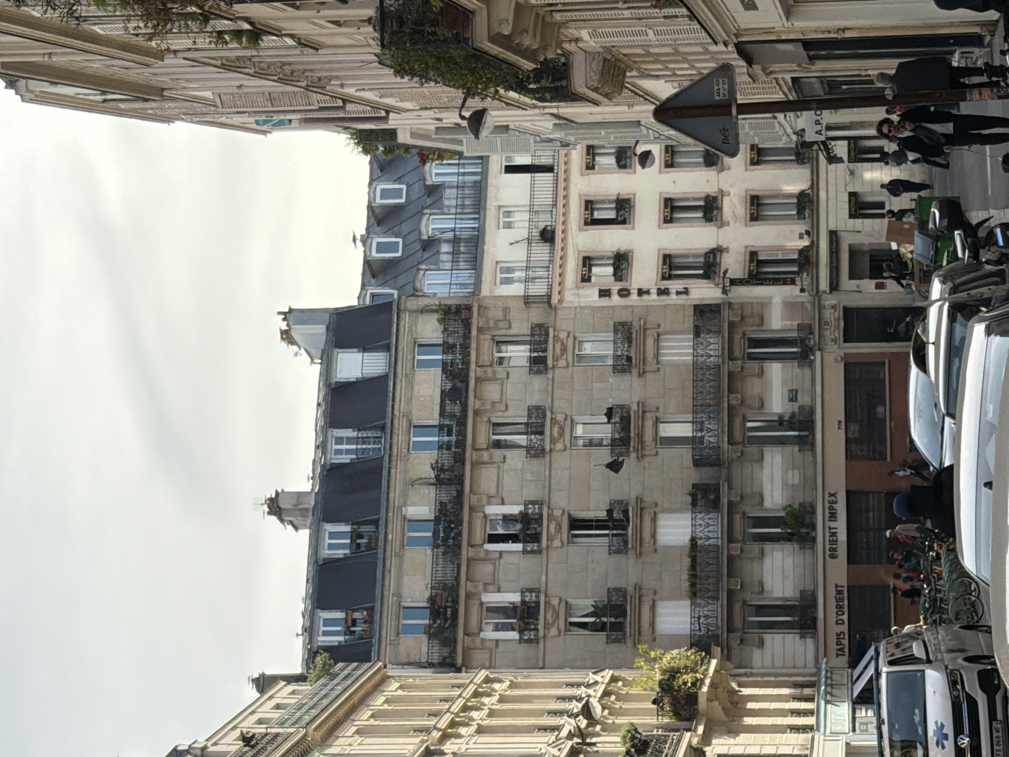This image captures a vertical street scene in Paris, characterized by its classic Haussmannian architecture under an overcast daytime sky. Multi-story buildings line both sides of the street, featuring ornate stone facades, numerous windows often adorned with wrought-iron balconies or railings, and distinctive gray mansard roofs with dormer windows. Greenery is visible in planters on some balconies and ledges.
The street below is busy with urban life. Parked vehicles, including cars, motorcycles, and scooters, line the curb. A white van, displaying a "Star of Life" symbol, suggesting it's an ambulance or medical transport, is also visible. Several people are present, walking on the sidewalks or standing near shop entrances. A prominent triangular traffic sign is seen on the right side of the street, alongside a lamppost.
Visible text on the buildings includes "TAPIS D'ORIENT" and "ORIENT IMPEX," indicating a rug store. Another building displays "HÔTEL MONNIER." The letters "A.P.C" are also seen on a storefront, likely belonging to a fashion brand. Other fragmented text includes "Garder," possibly part of "Garderobe," and a vehicle license plate reading "FX 068-WF." Further text fragments like "net," "69," "SP 27 91," and "Tommun" are present but their context is less clear. The scene depicts a typical, bustling street in the city, showcasing its architectural heritage and daily activity.