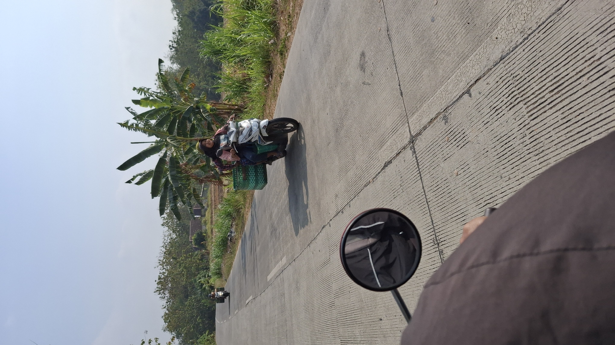 A woman wearing a headscarf and a pink shirt rides a white scooter on a paved road in Pucakwangi, Indonesia. She is carrying a large green woven basket filled with produce on the back of the scooter. The road is lined with green vegetation and banana trees. In the background, there are some buildings and fields. Another person on a scooter is visible further down the road. The image is taken from the perspective of someone riding behind her on another scooter, with a motorcycle's side mirror in the foreground.