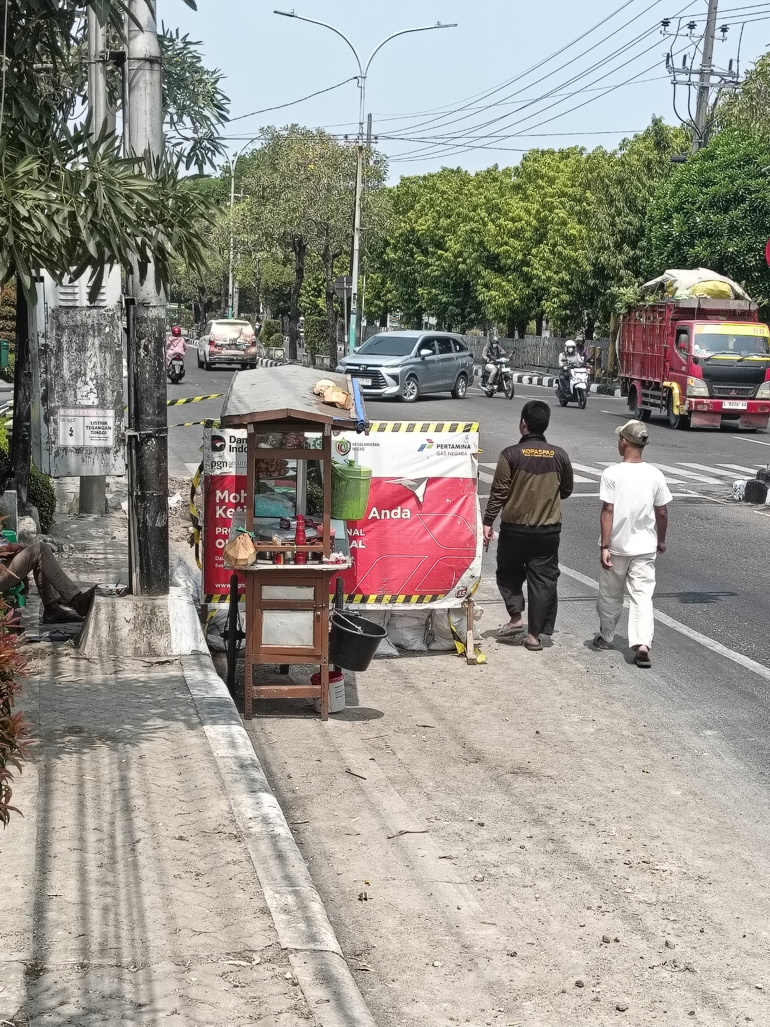 The image depicts a street scene in Gapurosukolilo, Indonesia, on a bright, sunny day. A two-lane road is visible, with traffic moving in both directions. On the right side of the road, a red truck with a tarp-covered load is stopped, and its license plate is partially visible. Two men are walking on the right side of the road, away from the camera, one wearing a white t-shirt and a cap, and the other a dark jacket with "KOPASPAD" text. A barrier with a red and white banner reading "PERTAMINA GAS NEGARA" is erected along the road.
On the left side of the road, a sidewalk is present, where a small, wooden food cart with items on display is set up. A person is seated on the sidewalk next to a grey bag. Behind the food cart and along the sidewalk, there are more barriers and construction materials. A utility pole with electrical wires runs along the left side of the street. Trees line the sides of the road, providing shade. In the background, other vehicles, including cars and motorcycles, are seen on the road. The overall scene suggests an active urban environment with ongoing construction or roadwork.