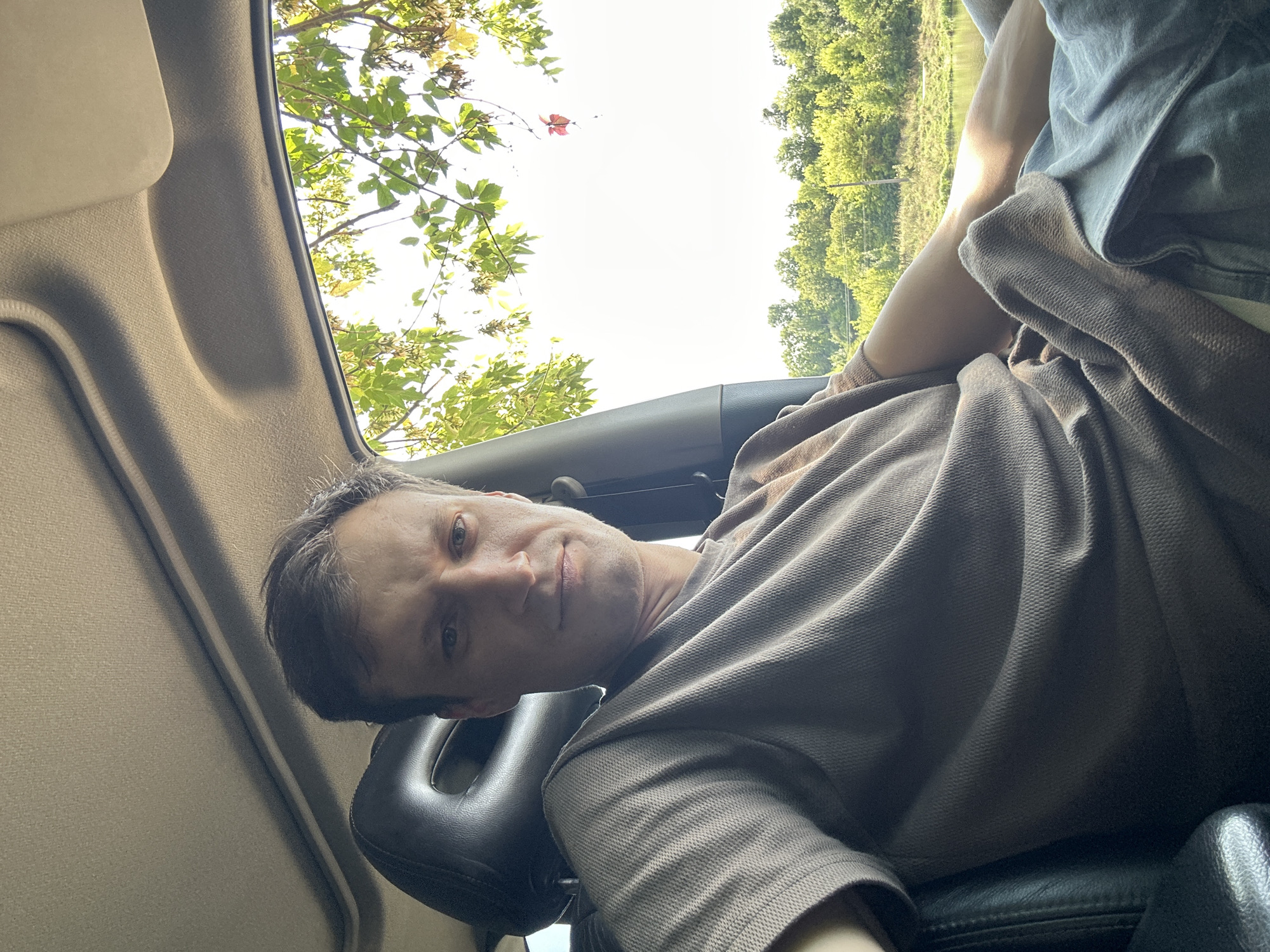 A man is captured in a selfie taken from inside a vehicle, presumably in or near the city of Makiv, Ukraine. The man, with short brown hair and a neutral expression, is wearing a brown, textured t-shirt. He is seated in the driver's seat, with the car's interior visible, including the headrest and part of the dashboard. Sunlight streams in from the right, illuminating the scene and casting a warm glow. Outside the car window, lush green trees and a body of water are visible, suggesting a rural or park-like setting on a clear day. The man's left arm rests on his lap, and his right arm is extended, holding the phone to take the picture. The overall impression is a candid moment of a person enjoying a scenic drive.