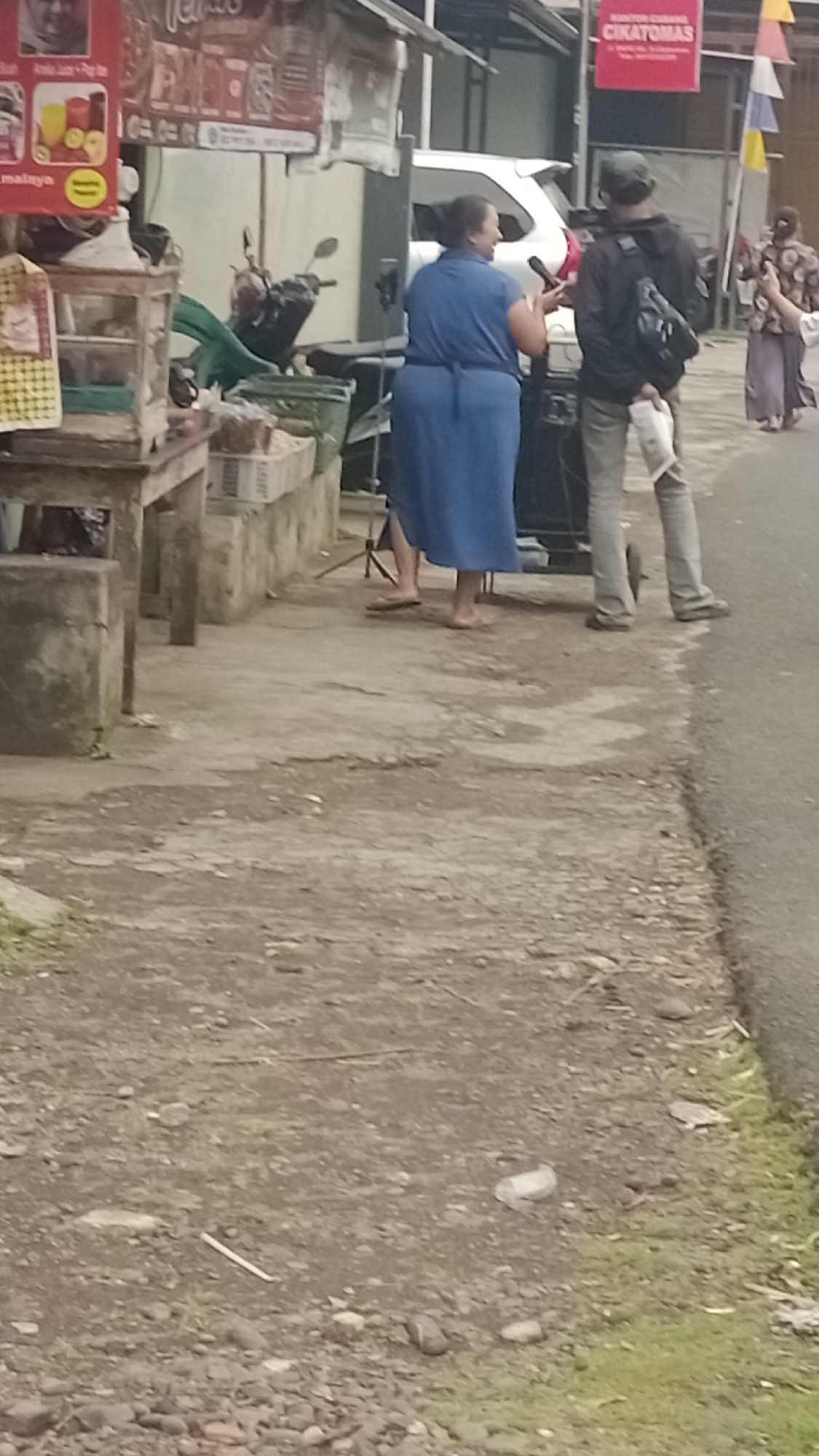 A blurry, medium-shot image taken outdoors captures a street scene. In the foreground, a dirt and gravel path leads towards a paved road on the right. A woman in a blue dress, facing left, is speaking into a microphone held by a person in a black hoodie and cap. The person in the hoodie is wearing grey pants and has a backpack. Behind them, a white car is partially visible. To the left of the woman, a wooden stall with glass display cases is set up, featuring various items, including what appear to be packaged snacks and some greenery. A motorcycle is parked near the stall. Further in the background, other individuals are walking, one wearing a patterned top and a long skirt. The scene suggests a casual outdoor event or market setting. The sign "CIKATOMAS" is visible on a red banner in the background. The image appears to be taken in Pakemitan, Indonesia.