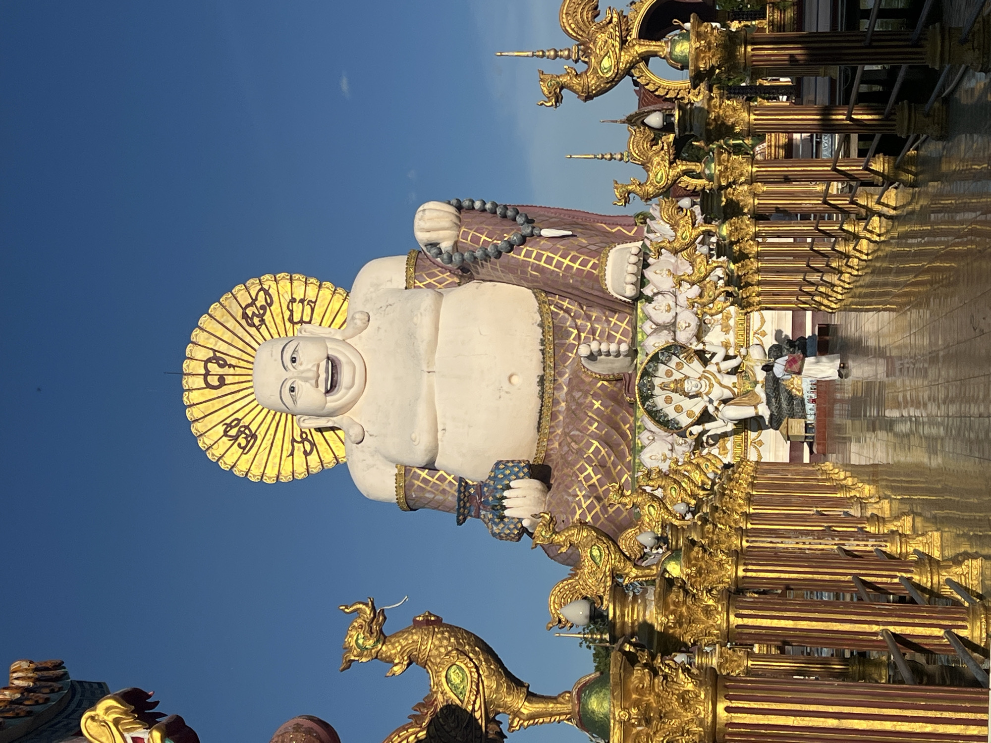 This media file features a large, cheerful statue of the "Laughing Buddha" in front of a clear blue sky. The statue is white, with a large belly and a radiant golden halo. It is adorned with a purple and gold patterned robe and a grey beaded necklace. To the right of the main statue, there are intricate golden pillars and decorative elements, some featuring mythical creatures, reflecting off a polished surface. Smaller figures are also visible behind the main statue and within the decorative structures. The overall scene is set outdoors, likely in a temple complex given the elaborate religious iconography and architectural style. The sunlight suggests it is daytime, possibly late afternoon given the long shadows. The image conveys a sense of joy and serenity associated with Buddhist imagery. The location is identified as Baan Plai Laem, Thailand, a known tourist destination with a prominent large Buddha statue.