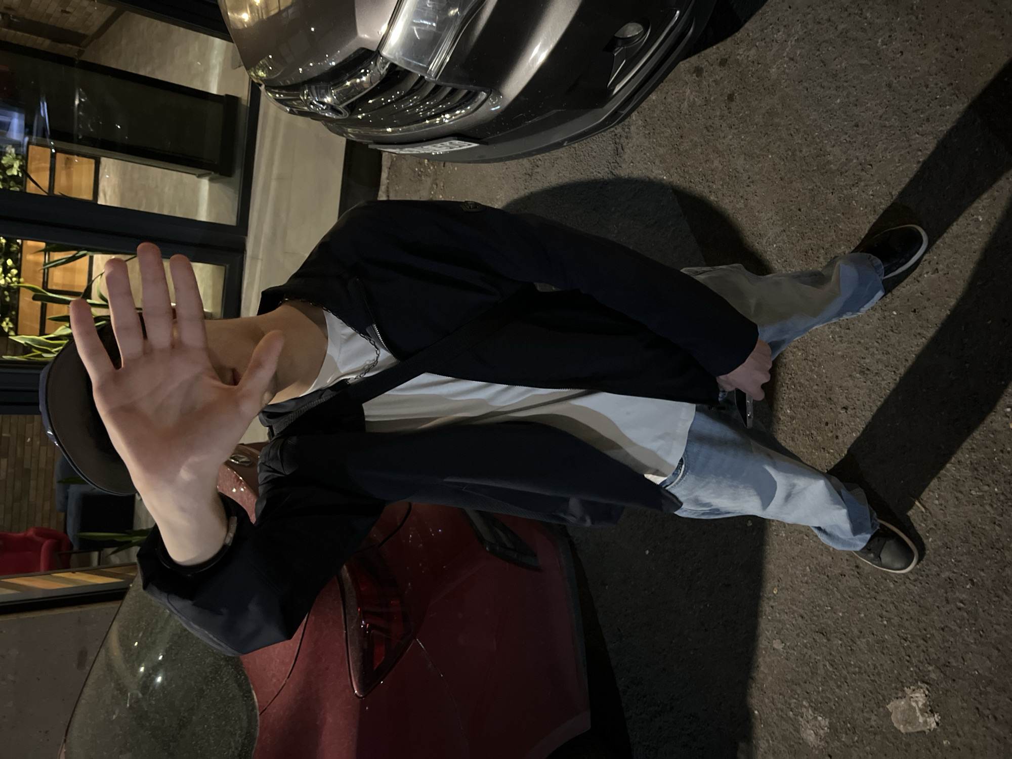 A person is standing in an urban environment near parked vehicles. The individual is wearing a dark jacket, white t-shirt, light blue jeans, and dark sneakers. The person has their hand raised, obscuring their face. The location appears to be a city street at night. Specific identification of Yerevan, Armenia, is not possible based solely on the image. There are two cars, one red and one grey, parked nearby.