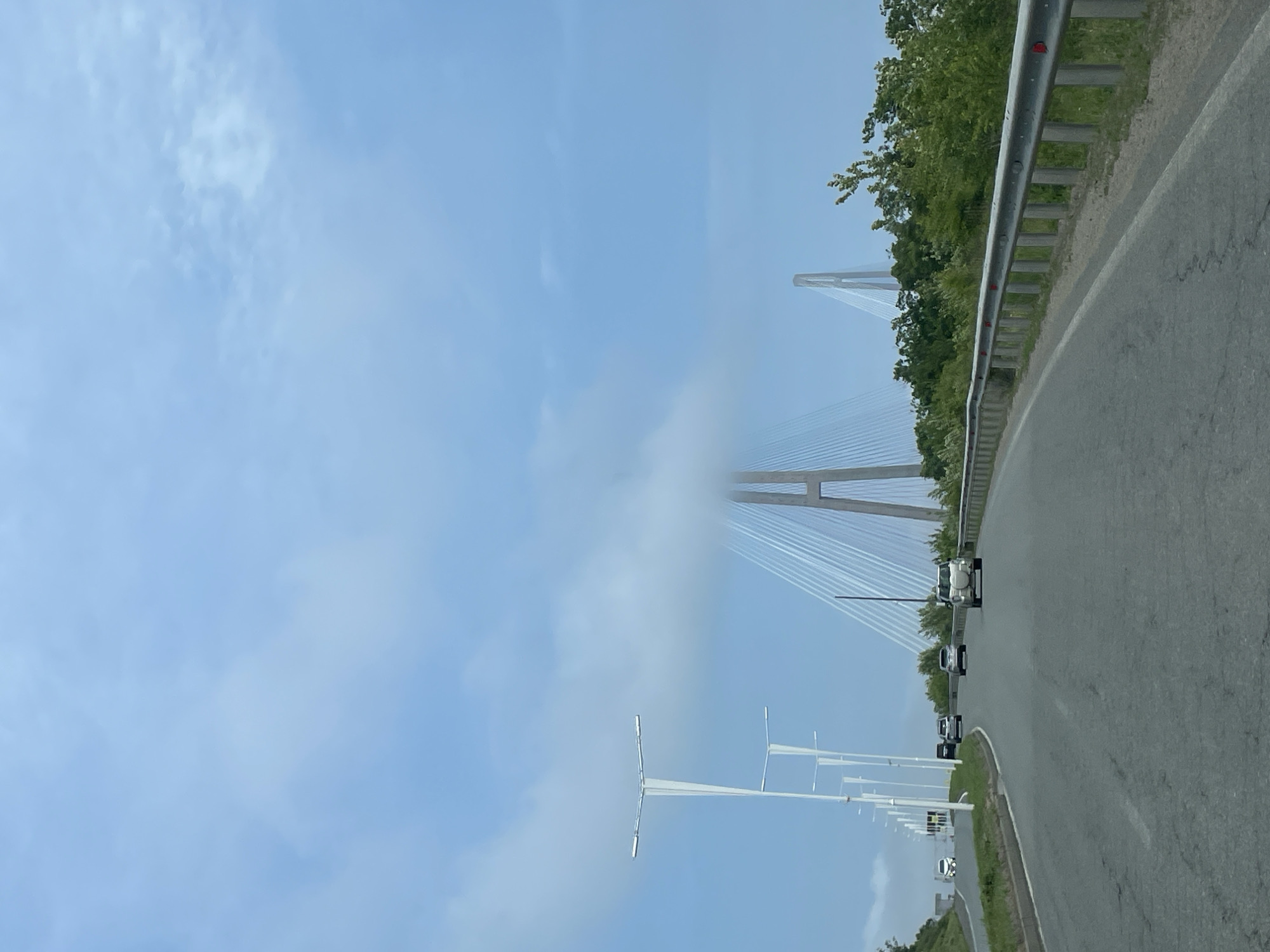 The image depicts a daytime scene of a highway approaching the Russky Bridge in Russky, Russia. The road, made of dark asphalt with a white dividing line, curves gently to the right, with several vehicles, including sedans and SUVs, traveling away from the viewer.
On the right side of the road, a metal guardrail with visible red reflectors runs parallel, backed by a dense line of green trees. To the left, a series of modern, white streetlights or utility poles line the road.
Dominating the mid-ground and background is the striking white structure of the cable-stayed Russky Bridge. Its tall, A-shaped towers and intricate network of support cables are visible, but a significant portion of the bridge's main span and lower tower sections are enveloped and partially obscured by a thick band of low-hanging clouds or fog, creating an atmospheric, almost surreal effect. Above this cloud layer, and in sections not covered by it, the sky is a clear, bright blue with scattered, lighter clouds. The weather appears to be a mix of clear skies and localized fog or low clouds around the bridge structure. No visible text is present in the image.