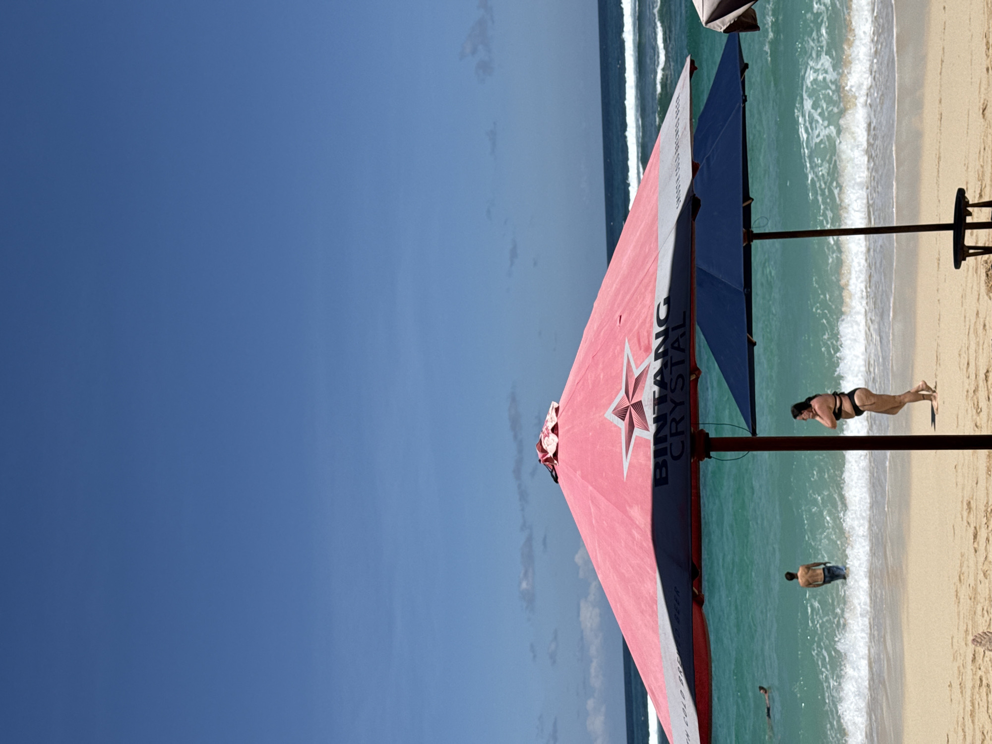 The image depicts a beach scene, likely in Kuta Selatan, Indonesia, given the Bintang Crystal umbrella, a popular beer brand in Indonesia. The beach is sandy with gentle waves breaking along the shoreline. Several people are visible: a woman in a black bikini doing yoga or stretching, a man standing in the shallow water, and at least one other person swimming further out. A large beach umbrella with a red and blue canopy featuring the Bintang Crystal logo dominates the foreground. The sky is clear and blue.