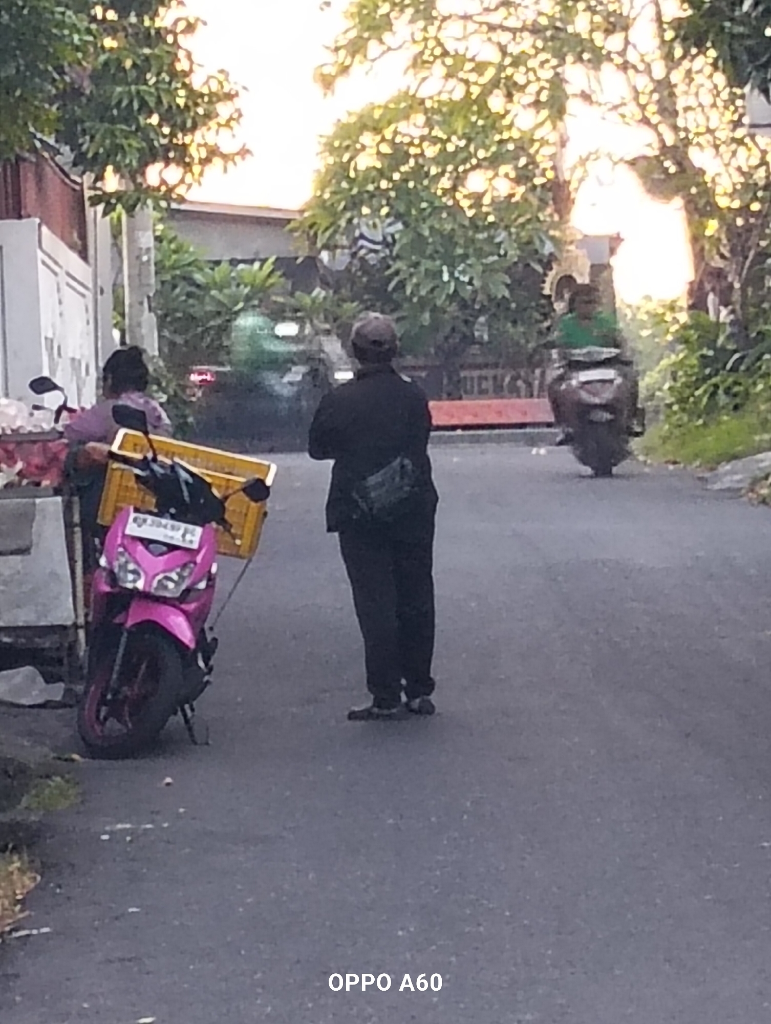The image depicts a street scene in North Kuta, Indonesia, likely during daylight hours, with trees and vegetation visible. In the foreground, a pink scooter is parked next to a vendor stall. A person in a purple top is standing behind the stall, attending to what appears to be produce. A yellow crate is mounted on the back of the scooter. Further down the street, another person is walking away from the camera, wearing dark clothing and a black bag slung across their body. In the background, a person on a dark-colored motorcycle is moving away from the viewer. The overall atmosphere is that of a typical day in a bustling Indonesian town, with elements of commerce and transportation present.