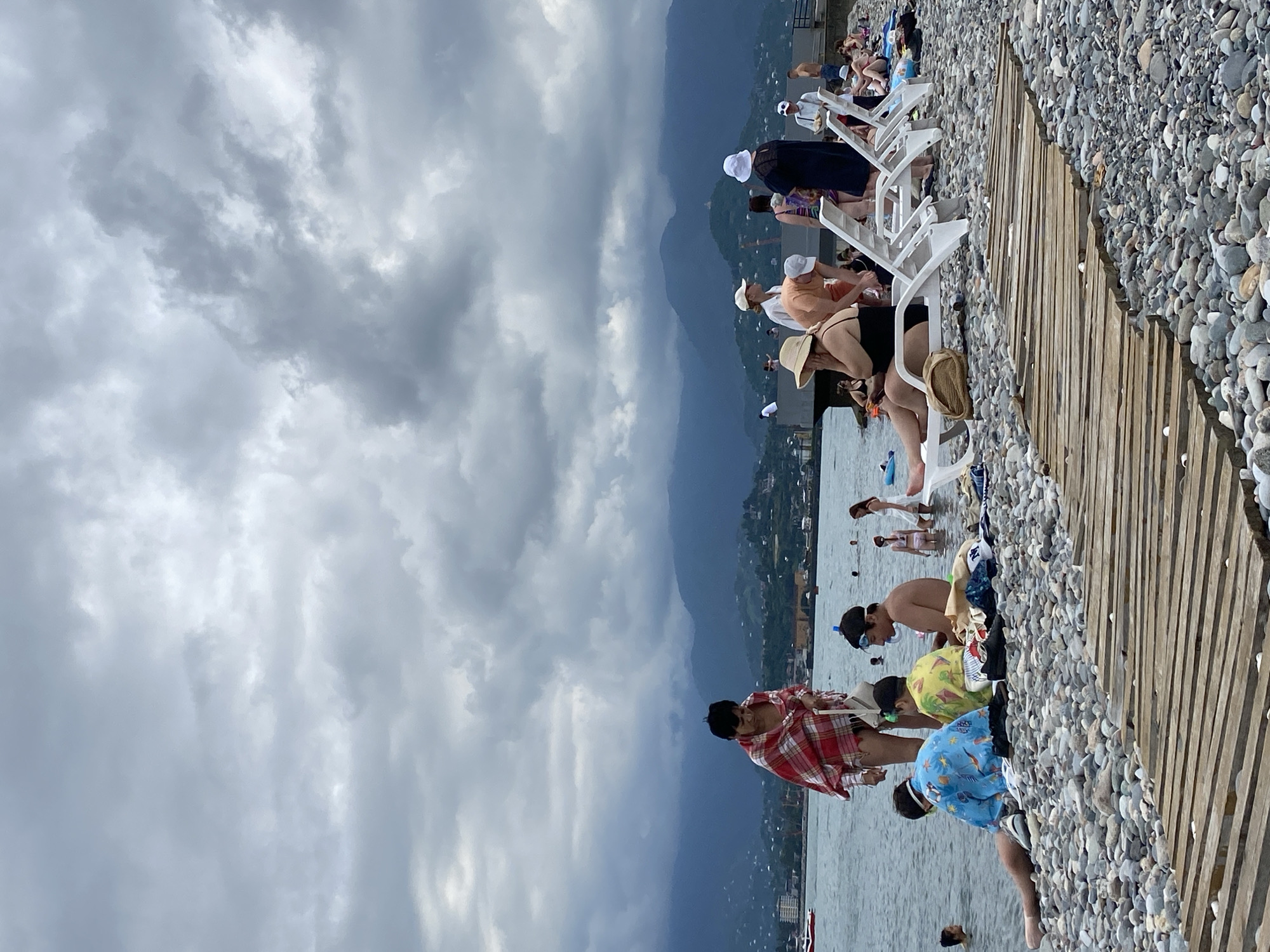 The image depicts a crowded pebble beach in Batumi, Georgia, with a cloudy sky and mountains in the background. In the foreground, a wooden path is laid on the pebbles. Several groups of people are visible on the beach. On the left side, near the water, a person wrapped in a red and white checkered blanket stands near the water's edge. In front of them, two children wearing swim trunks and goggles are sitting on the pebbles. To the right, several people are relaxing on white lounge chairs. Some are wearing hats and swimwear. Further back, more people are seen in the water and on the beach. The sea is calm, and a few boats are visible in the distance. The overall atmosphere appears to be a typical summer day at the beach with a significant number of visitors.
