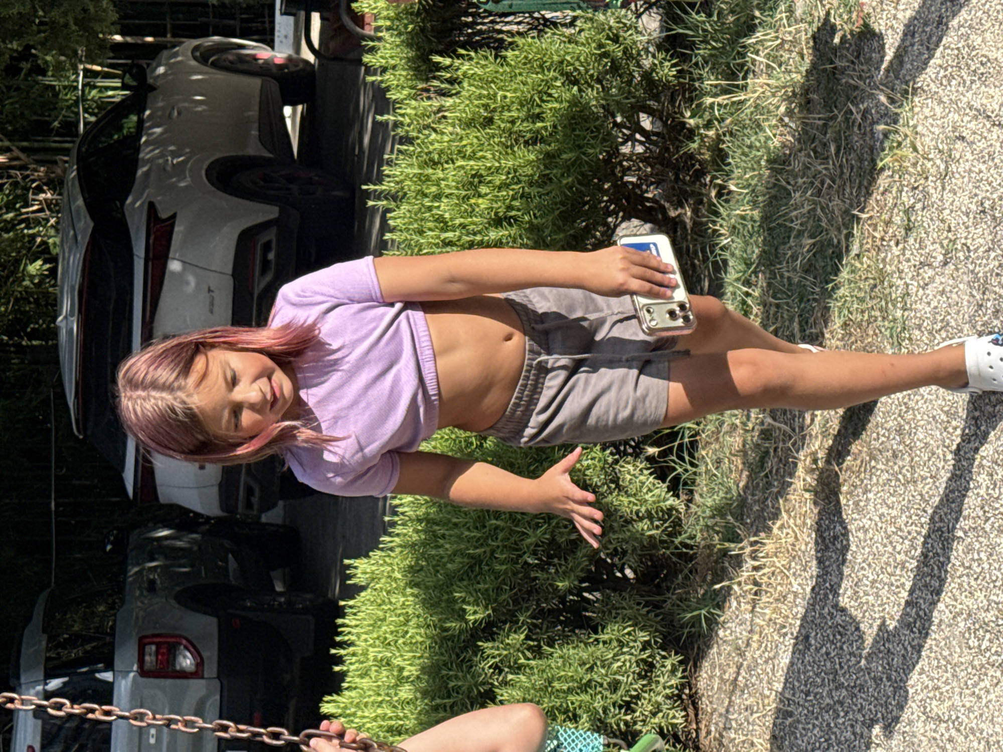 In Partenit, Ukraine, a young girl with pink-tinged hair walks outdoors, holding a white mobile phone in her right hand. She is wearing a lavender crop top and grey shorts. The background features dense green bushes and two parked vehicles, a light-colored SUV and a darker SUV, suggesting a residential or public area. The ground is covered in gravel and dappled with sunlight and shadows.