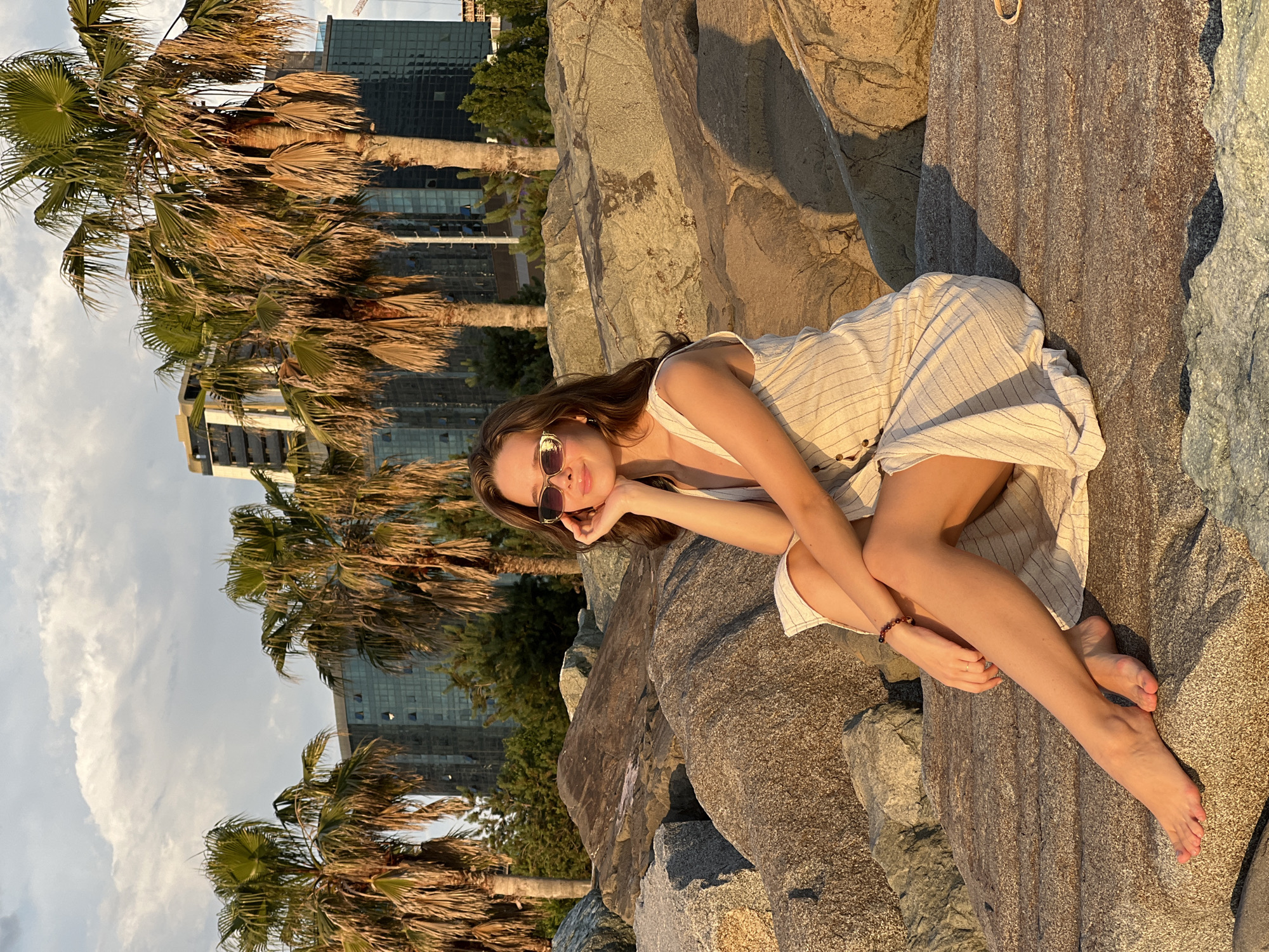 In Batumi, Georgia, a young woman sits on large rocks along the coastline, bathed in the golden light of a sunset. She is wearing a light-colored, pinstriped dress and sunglasses, with one hand resting on her chin. Behind her, palm trees sway, their fronds silhouetted against a sky filled with soft clouds. In the distance, modern buildings with glass facades rise, reflecting the warm hues of the setting sun. The scene is serene, with the natural beauty of the coastline meeting the urban landscape.