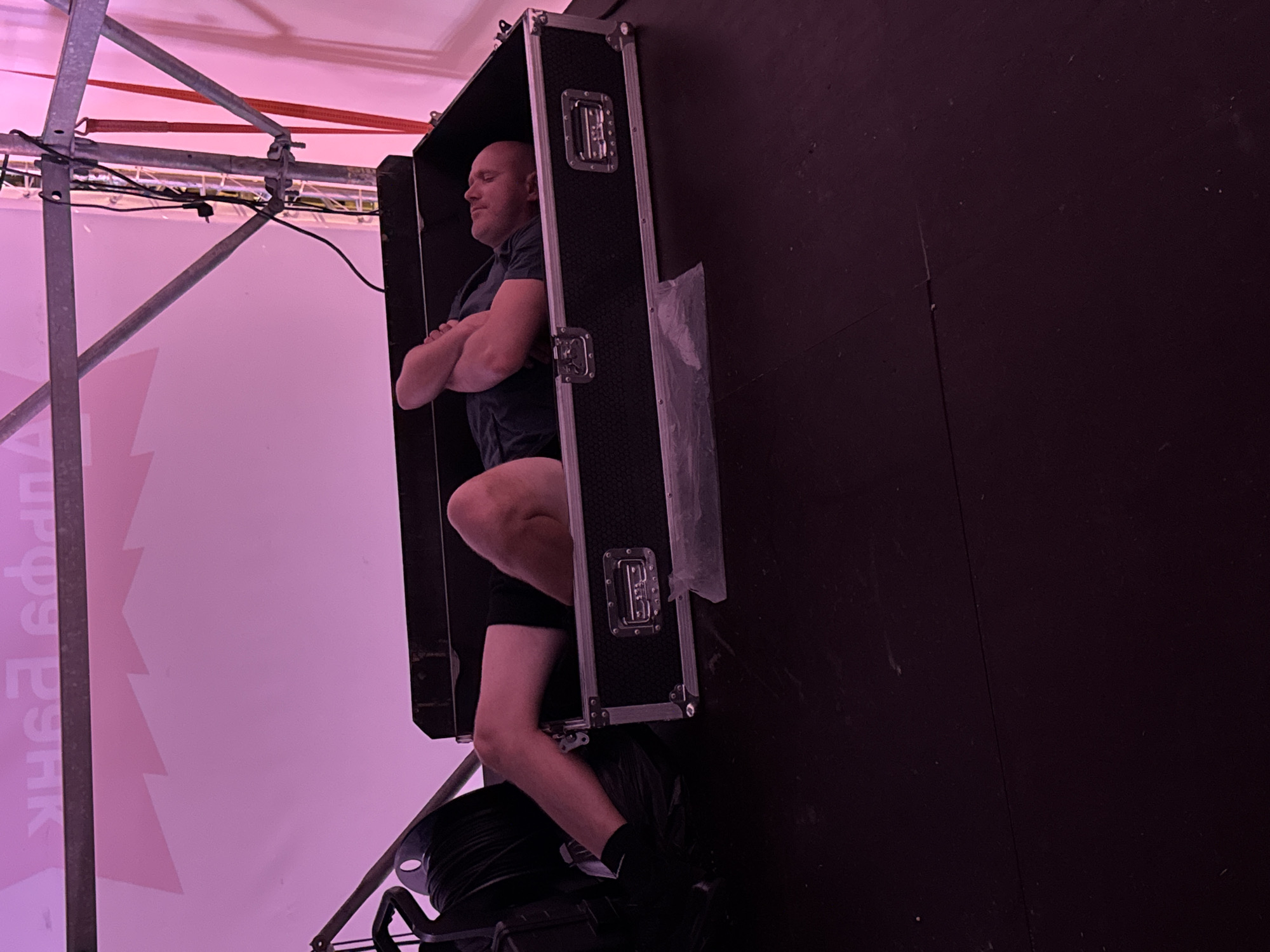 A bald, fair-skinned man with folded arms is lying down inside an open black road case. The case has silver latches and reinforced corners. The man is wearing a dark t-shirt and dark shorts. His legs are bent and extended out of the case. His feet are bare and crossed at the ankles. The road case is on a dark, flat surface. In the background, a pink banner with Cyrillic lettering is visible, as is scaffolding and lighting equipment. The environment appears to be an indoor or covered outdoor venue.