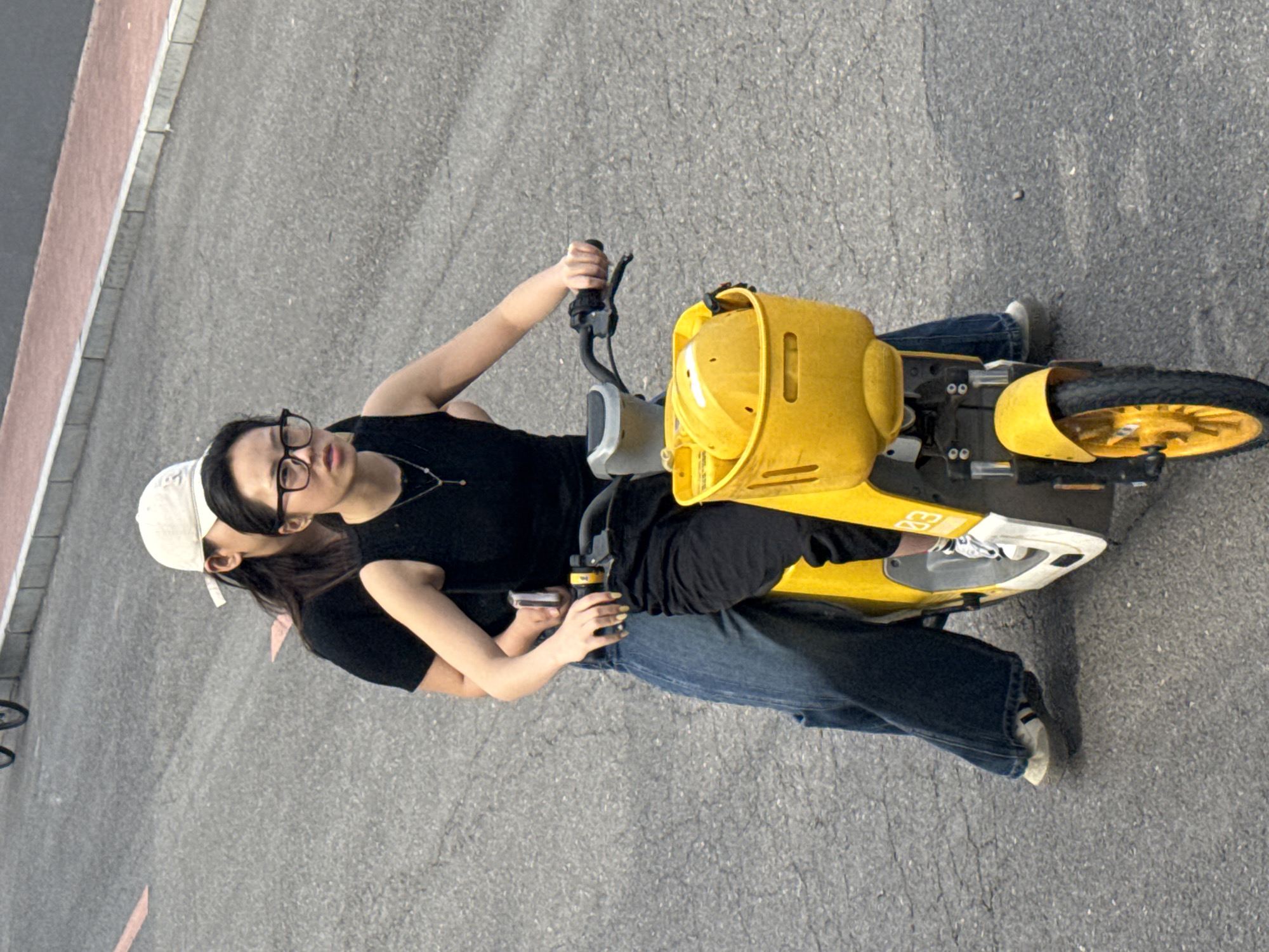 An overhead shot captures two individuals on a yellow electric scooter on an asphalt surface, likely a road or paved area in Zhongtun, China. The image is taken from a high vantage point, looking down at the subjects and their surroundings.
The person in the front, appearing to be the driver, wears a black sleeveless top, glasses, and blue jeans. Their right hand grips the scooter's handlebar. This individual has dark hair and black nail polish. The second person, riding as a passenger behind, wears a white baseball cap, a black t-shirt, and blue jeans. The passenger's left hand is visible, holding a smartphone. Their white sneakers with black stripes are also visible.
The scooter itself is a bright, conspicuous yellow, featuring a large front section that may serve as a basket or protective casing. It has a white and gray foot platform where the riders' feet rest. A small black number "93" is visible on the side of this platform. The background consists of dark gray asphalt, with a light gray curb and a reddish-brown strip visible along the left edge of the frame, suggesting the presence of a sidewalk or pedestrian area. The lighting indicates it is daytime with clear weather. The overall scene suggests a casual moment of transit or leisure.