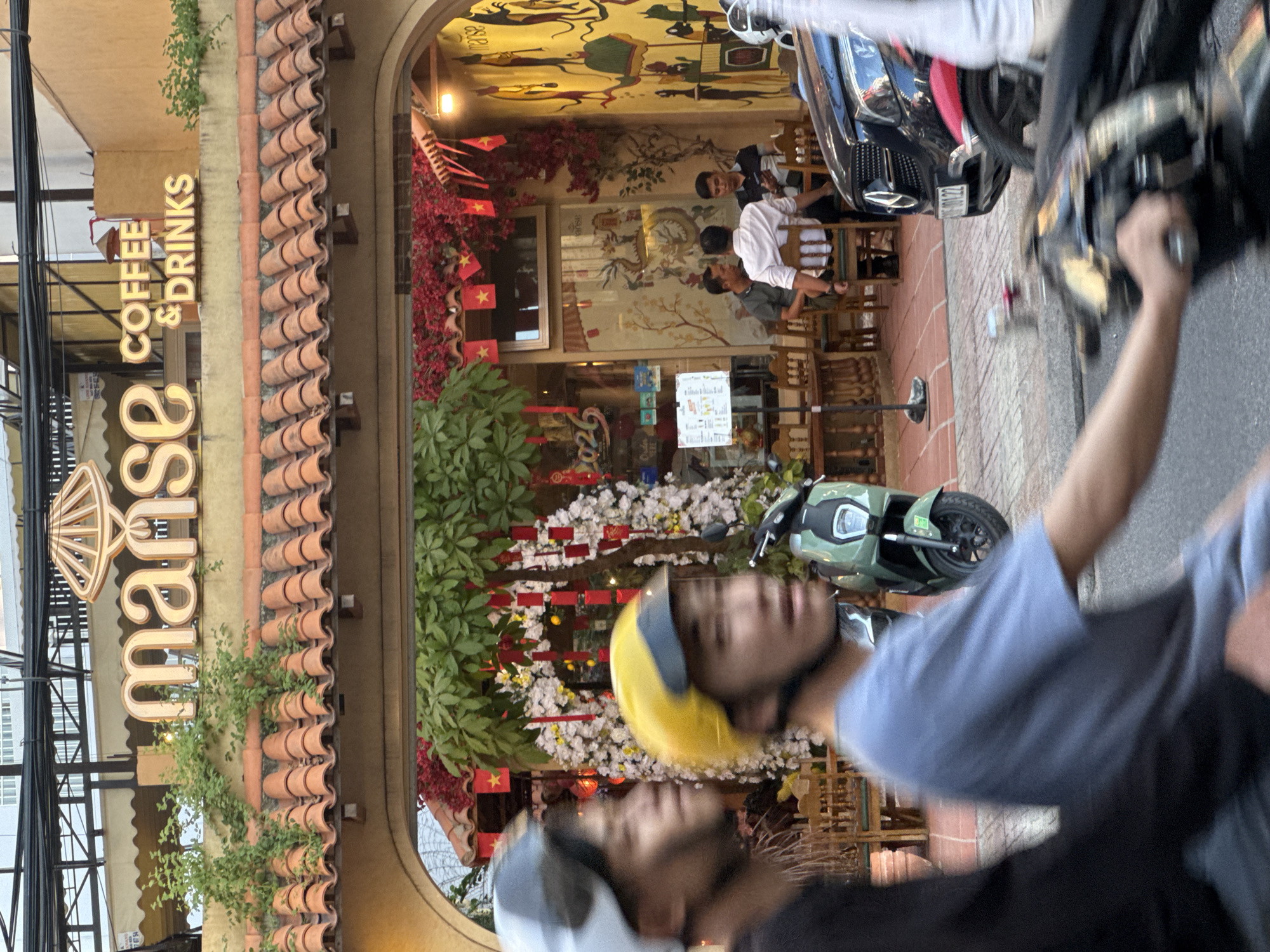 The image captures the exterior of a cafe named "manse COFFEE & DRINKS" located in Nha Trang Ward, Vietnam. The cafe features a terracotta-tiled roof and a vibrant facade adorned with red flowers and small Vietnamese flags. Inside, several individuals are seated at tables, some engaging in conversation. A green electric scooter is parked outside, and two individuals wearing helmets are riding on a motorbike in the foreground, their faces blurred due to motion. The overall scene suggests a bustling street-level cafe environment in a Vietnamese city.