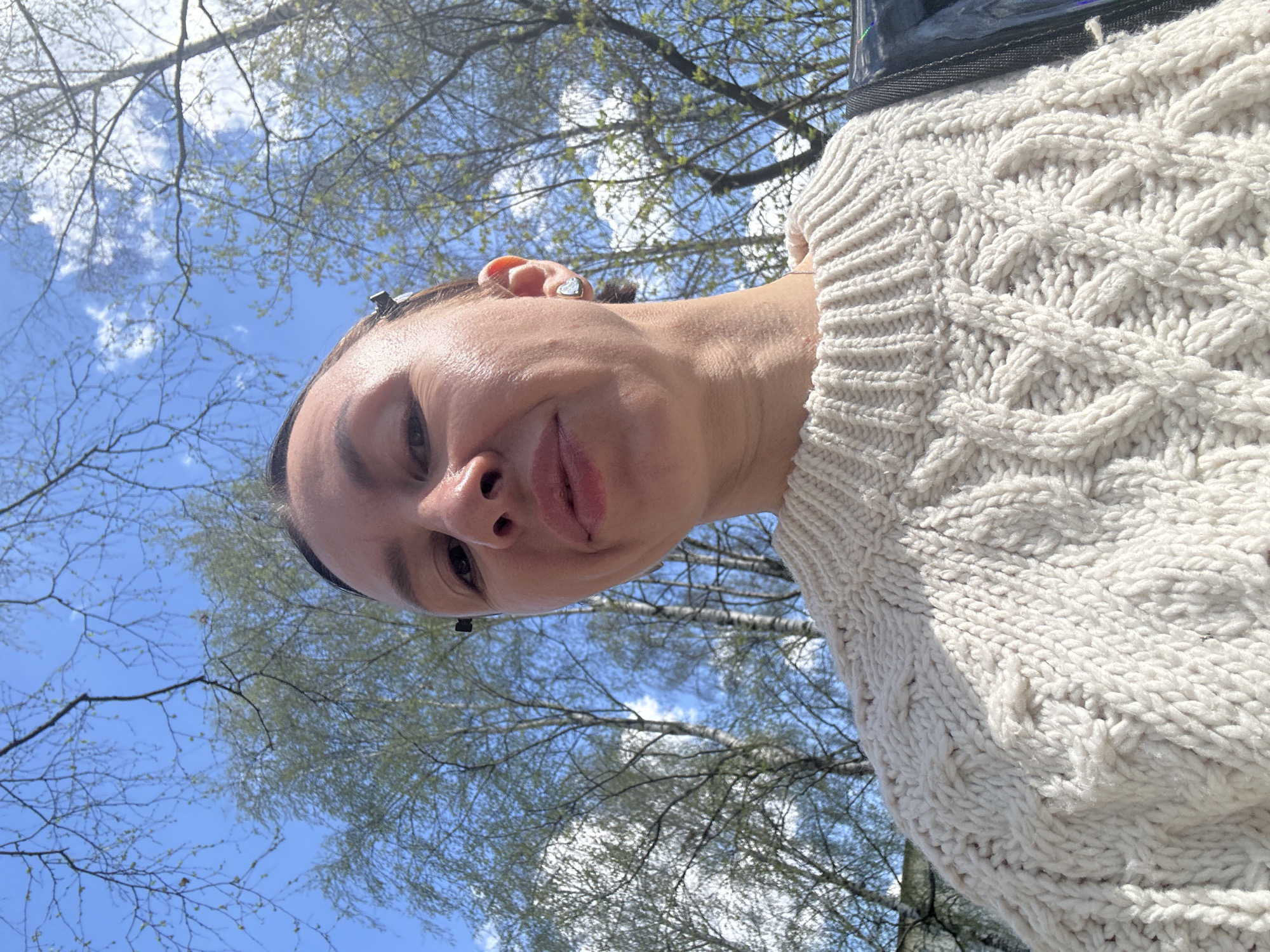 The image is a selfie taken from a low angle, looking up at a woman wearing a thick, cream-colored cable-knit sweater. She is looking directly at the camera with a slight smile. The background shows a blue sky partially obscured by the bare branches of trees with small, budding leaves. It's daytime and the light is bright. The location cannot be determined to be Votkinsk, Russia, from the visual data provided.