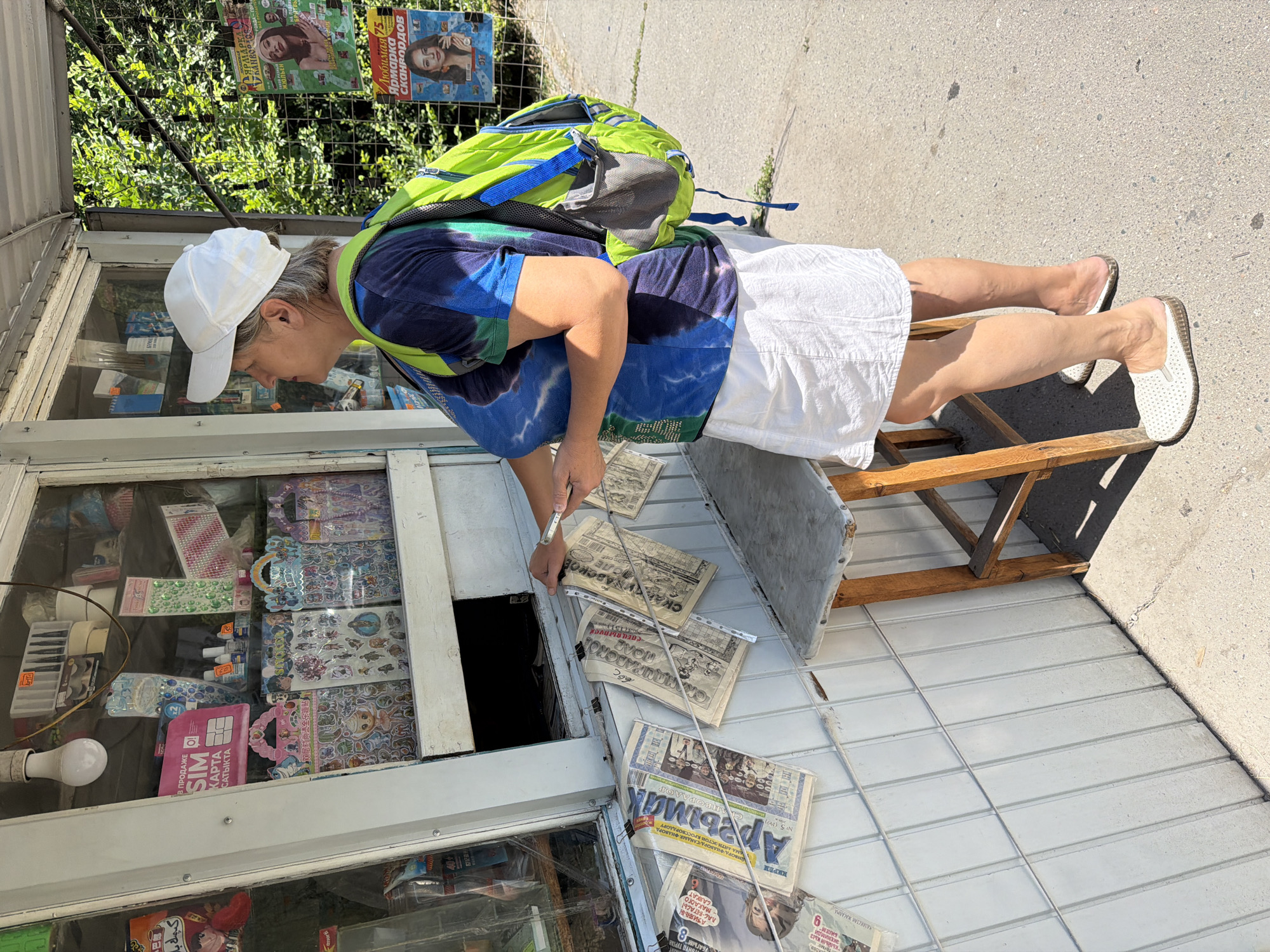 A woman wearing a white baseball cap, a blue and green tie-dye t-shirt, a white skirt, and white slip-on shoes stands on a sidewalk next to a kiosk. She is looking into the kiosk and holding a small metallic object, possibly a card or ticket, in her right hand. A bright green backpack with blue accents is slung over her shoulders. Newspapers are displayed hanging from clips on the side of the kiosk. The kiosk has a glass display window filled with various items, including stickers, SIM cards, and other small goods. A wooden stool with a worn top is positioned next to the kiosk. The environment appears to be outdoors, with trees and foliage visible in the background. The ground is paved with concrete.
