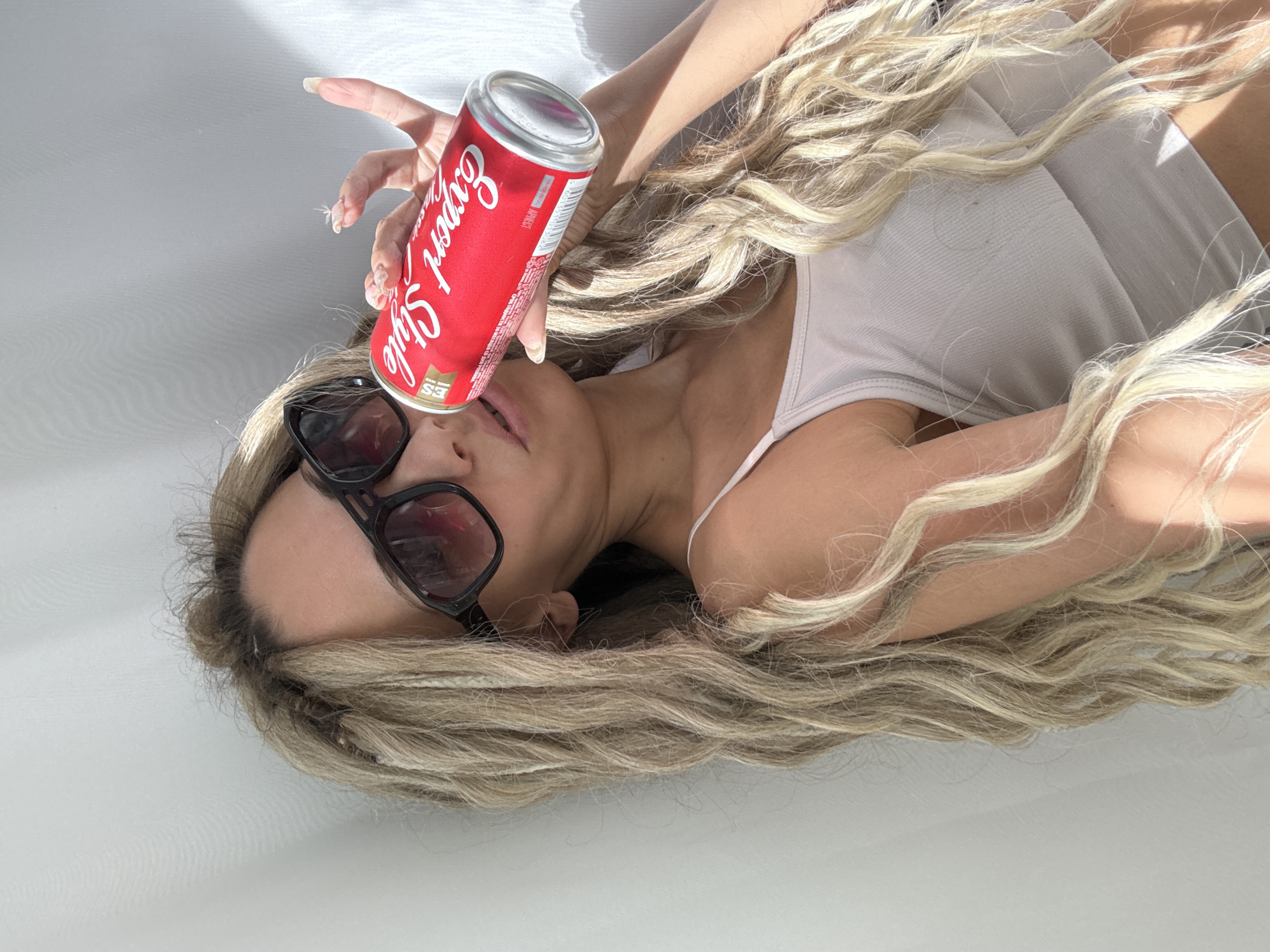 The image displays a person, likely female, lying down against a plain, light-colored background, possibly a white floor or bed. Their head is angled to the left, and long, voluminous wavy blonde hair spreads around their head and shoulders. The person is wearing a light-colored (possibly off-white or beige) ribbed tank top and black-framed sunglasses, which obscure their eyes.
A red aluminum can is held in the person's right hand, positioned above their mouth and tilted downwards, as if about to pour its contents. The can features prominent white cursive text that reads "Expert Style," with smaller text below it stating "The Natural" and "SINCE 1950," alongside an "ES" logo. The top of the can also shows "26.04" and the Russian word "АПЕЛСИН" (Apelsin), meaning "Orange."
The scene is brightly lit, likely by natural sunlight entering an indoor space, creating sharp, distinct shadows on the background and across the person's body and hair. This suggests it is daytime, possibly morning or afternoon. The activity appears to be a posed moment, perhaps for a beverage advertisement or a casual photo. The visible Russian text on the can is consistent with the provided location context of Rusanovo, Russia.