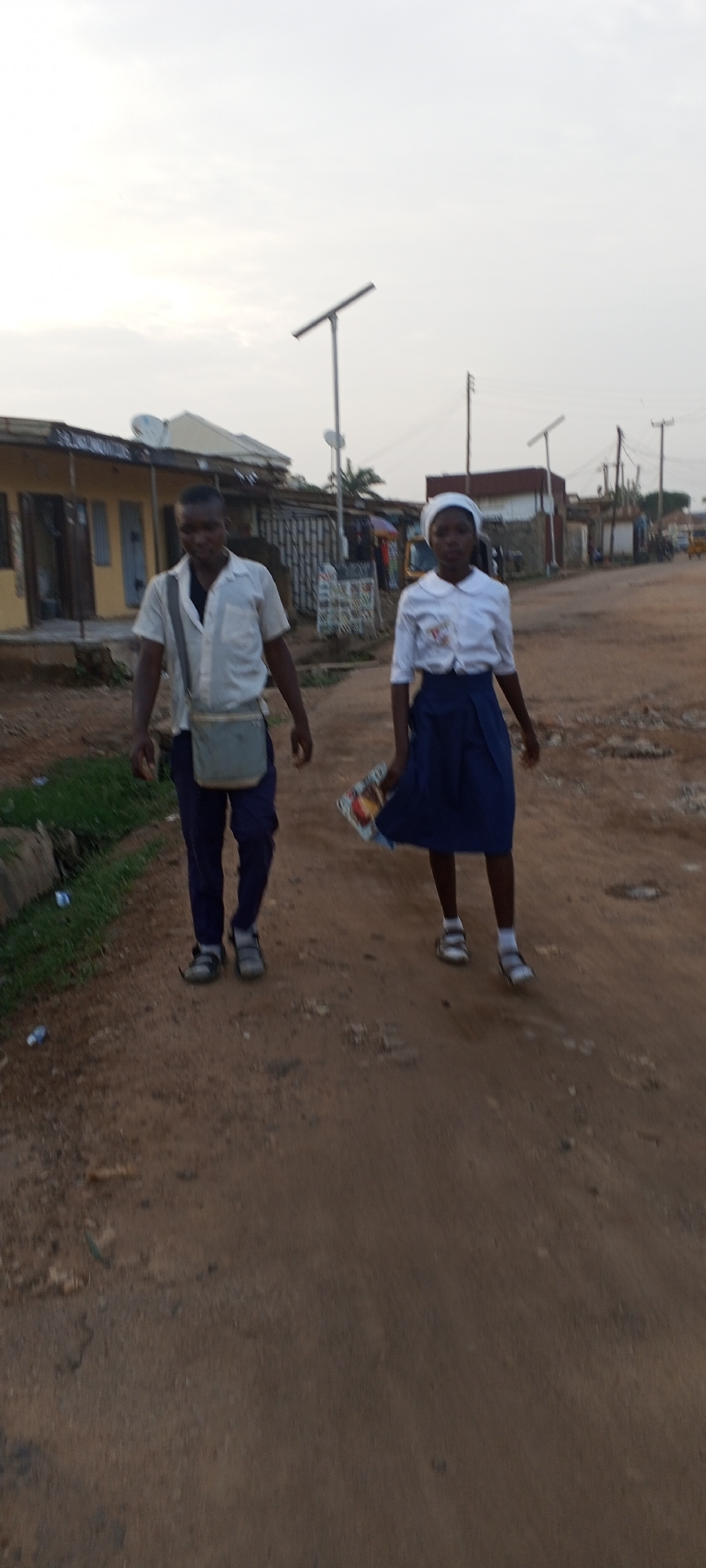 The image shows a man and a young woman walking on a dirt road in Sabon Tasha, Nigeria. The man is wearing a light-colored short-sleeved shirt, dark pants, and carries a satchel across his body. The young woman wears a white short-sleeved shirt, a dark blue skirt, white socks, and sandals. She is carrying some papers in her hand. The background shows buildings and utility poles, including solar-powered street lights. The sky is overcast. The ground is rough and unpaved.