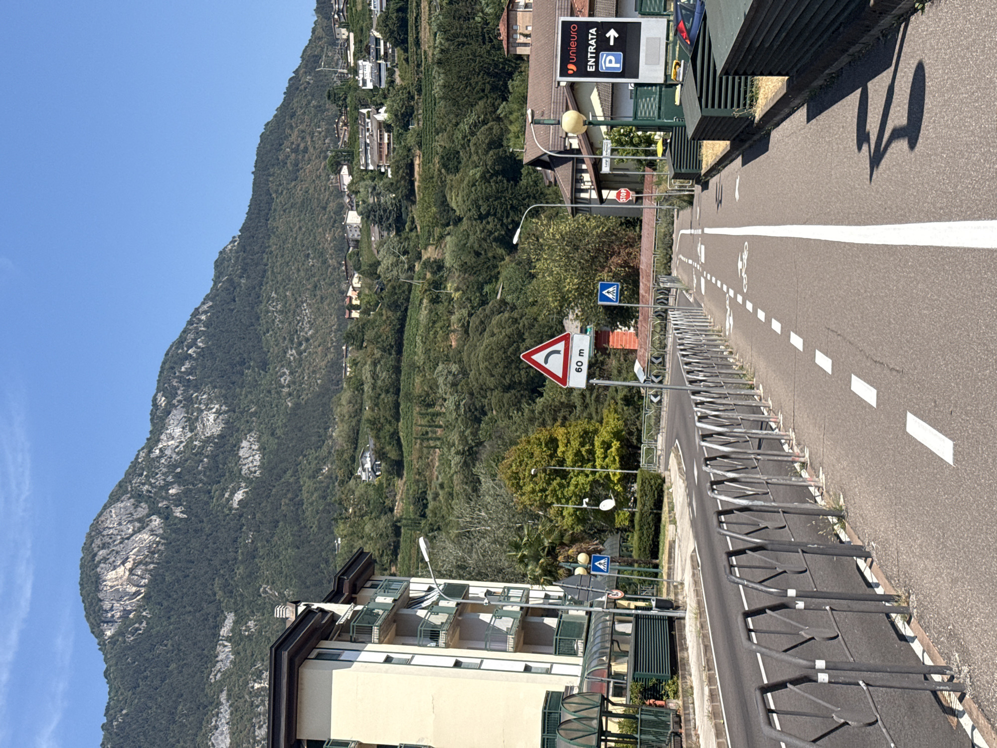 The image captures a street scene in Trento, Italy, on a clear, sunny day. The foreground features a paved road with a dedicated bicycle lane marked by white lines and bicycle symbols. A silver metal barrier separates the bicycle lane from the main road, which is also marked with white lines. In the background, a multi-story building with a cream-colored facade and green balconies stands to the left. To the right, a sign for "unieuro ENTRATA P" with an arrow indicates an entrance and parking. Further in the background, the lush green slopes of a mountain rise steeply, dotted with houses and vineyards. Traffic signs, including a curve warning sign and a pedestrian crossing sign, are visible along the roadside. The overall scene depicts an urban environment integrated with natural mountainous scenery.
